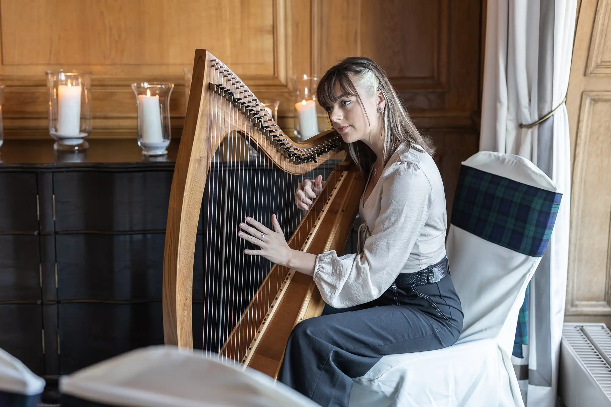 A woman wearing a light-colored blouse and dark pants, playing a harp in a room with wooden walls and lit candles on a cabinet.