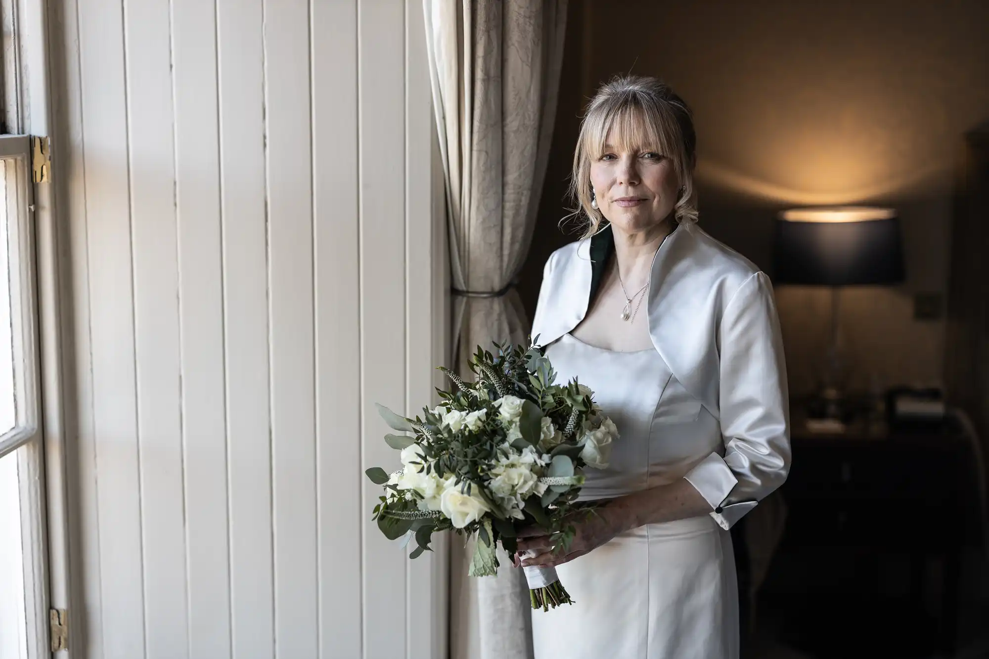 A woman in a white gown stands by a window holding a bouquet of white flowers and greenery, with a lighted lamp and a curtain in the background.