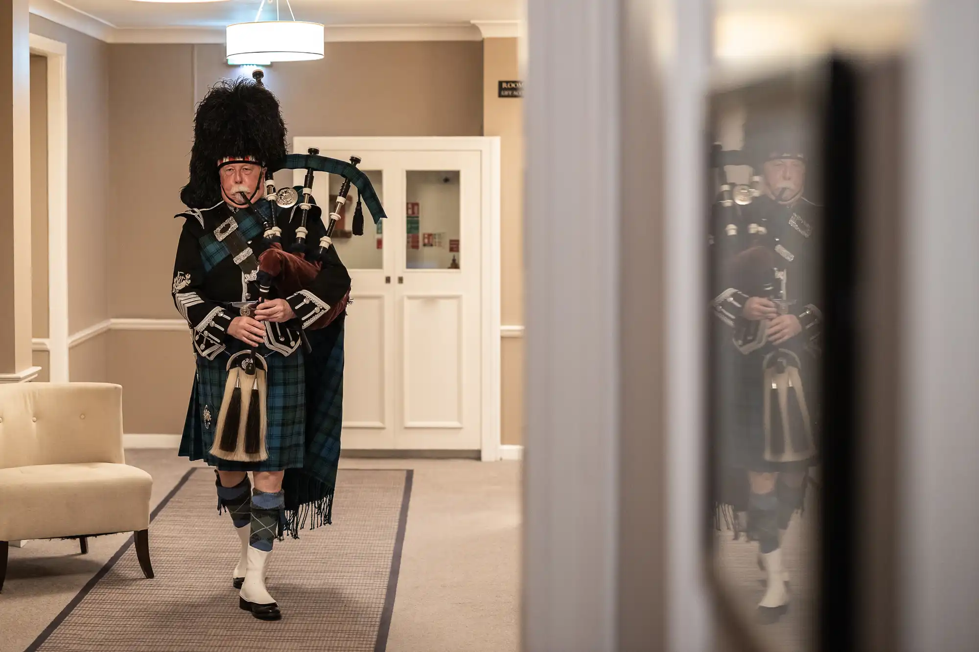 A person dressed in traditional Scottish attire plays the bagpipes while walking down a hallway beside a white wall and beige couch.