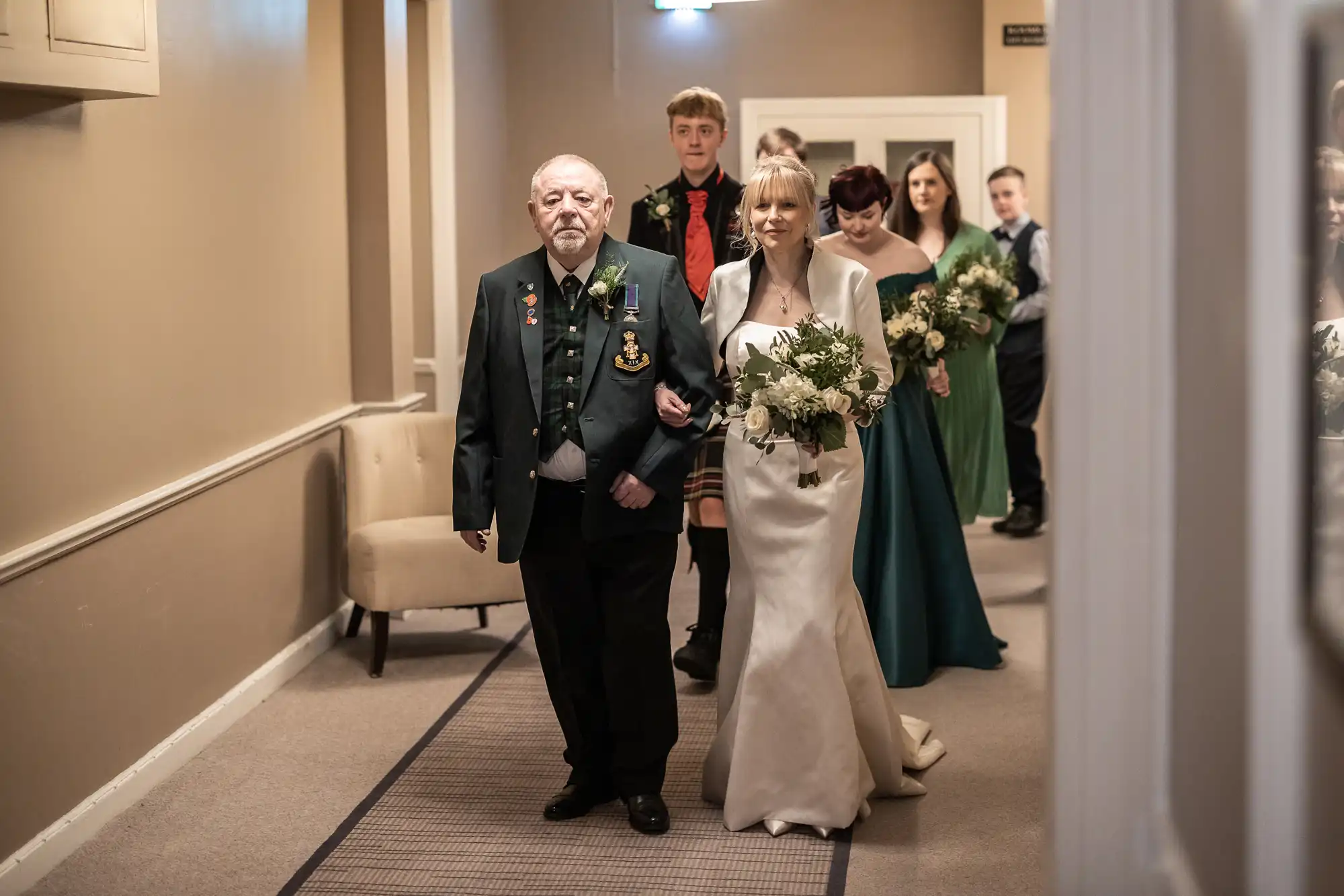 A woman in a white dress is escorted down a hallway by an elderly man wearing a green jacket. Bridesmaids holding bouquets and other people follow behind them.