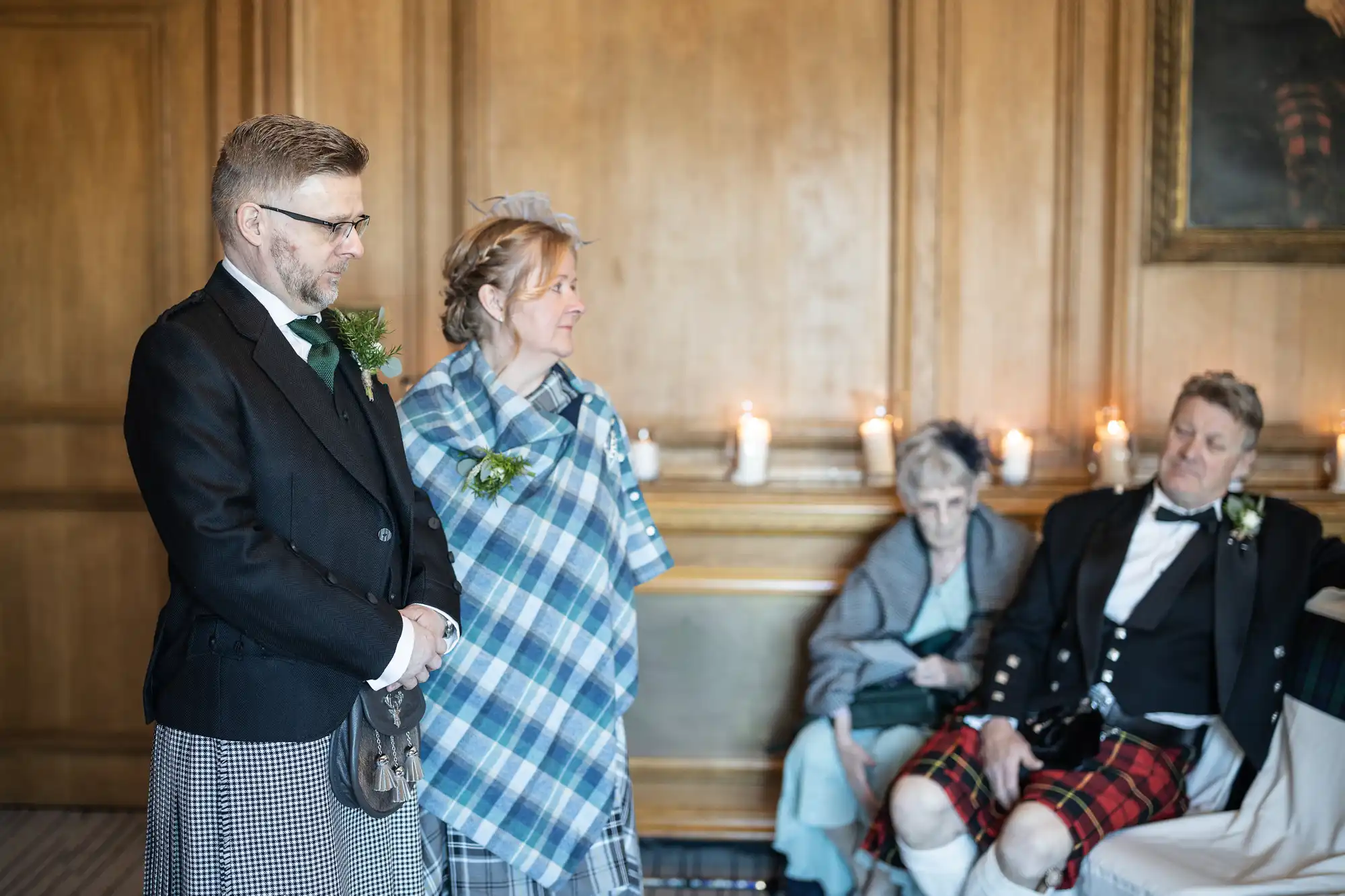 A couple stands dressed in traditional Scottish attire, while two older people sit in the background near candles on a wooden shelf.