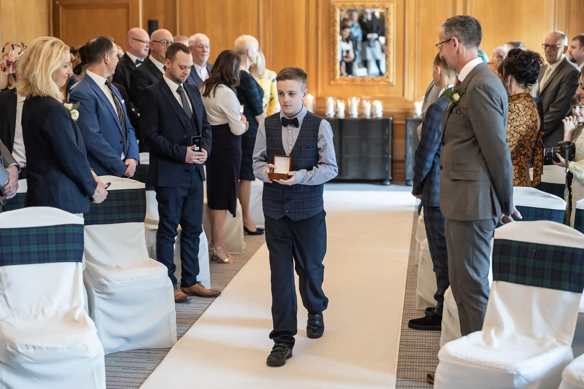 A young boy, dressed in a vest and bow tie, walks down the aisle holding a ring box, surrounded by seated and standing adults in formal attire at an indoor event.