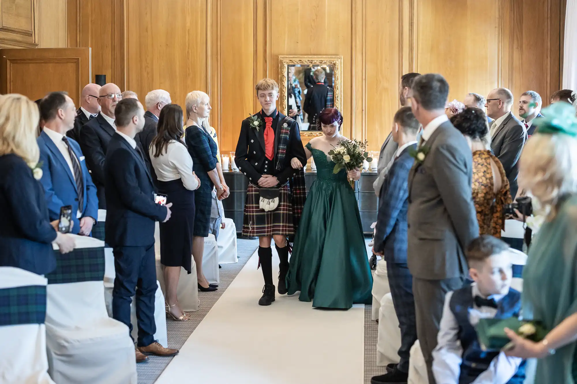 A man in a kilt escorts a woman in a green dress down the aisle at a wedding ceremony as guests seated on both sides look on.