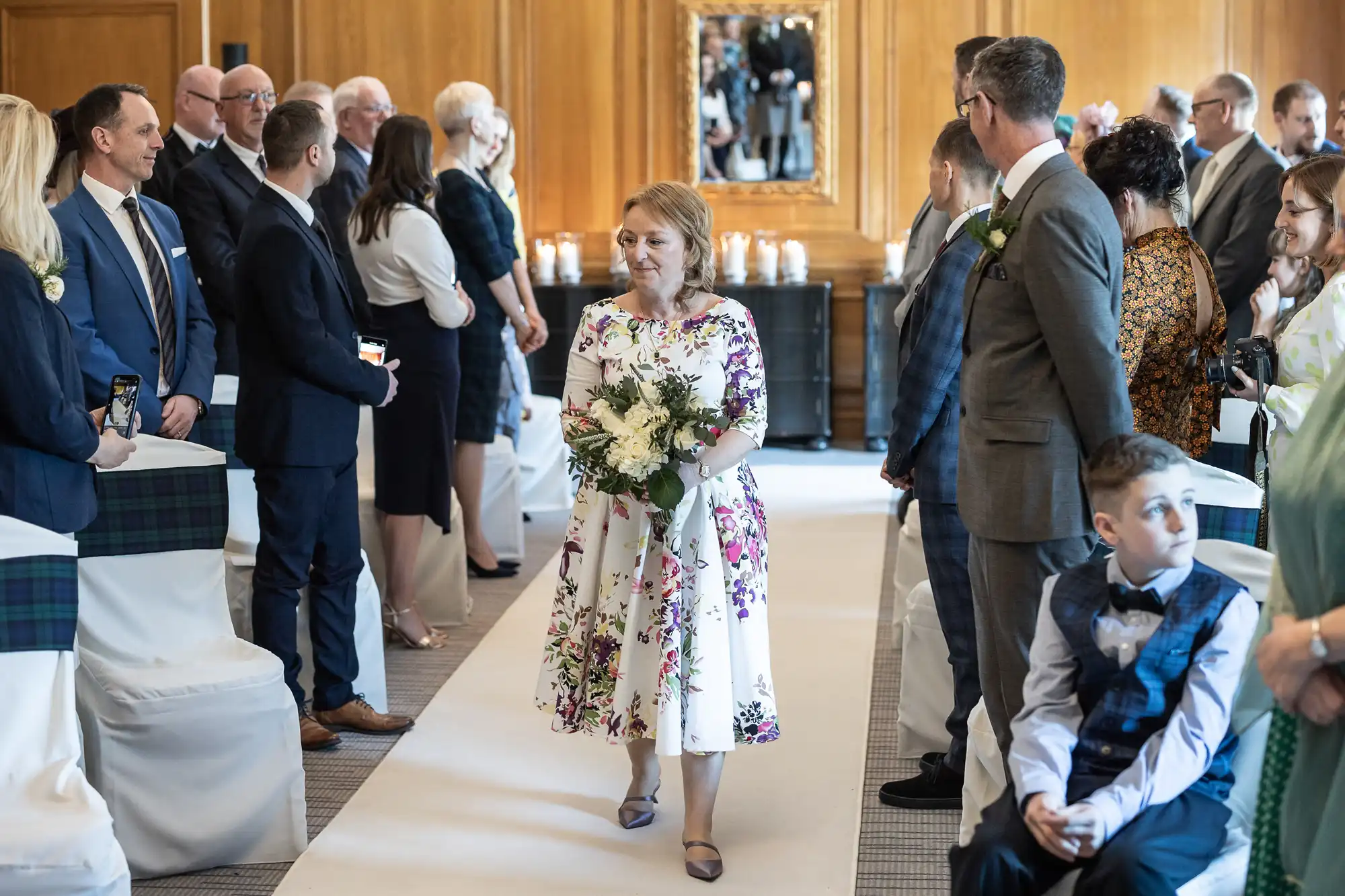 A woman in a floral dress walks down the aisle at a formal event, holding a bouquet, while guests seated on both sides watch.