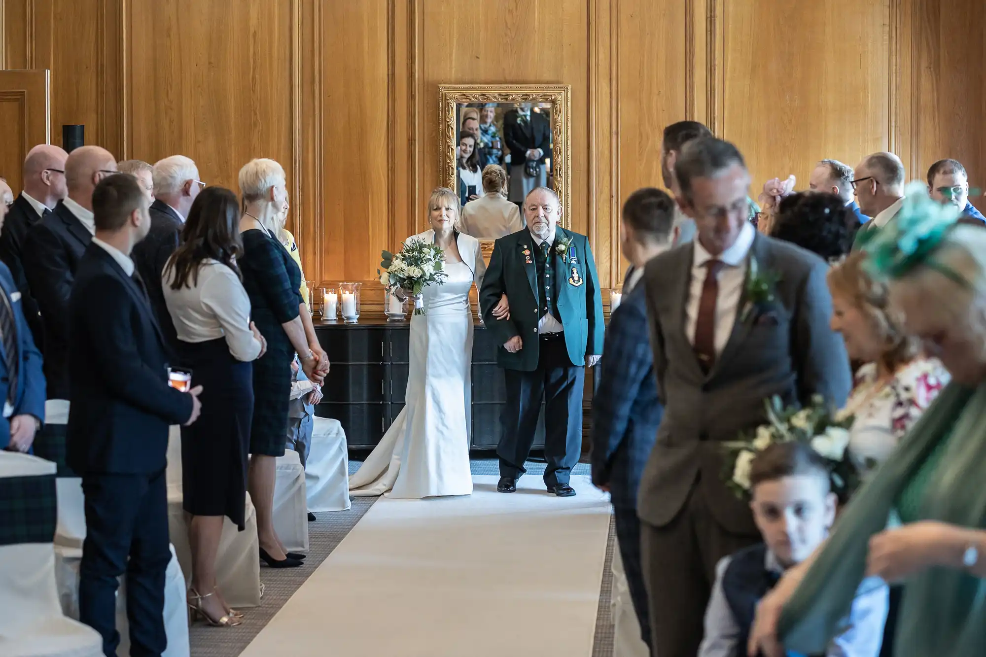 Bride in a white dress walks down the aisle with an elderly man in a green jacket, accompanied by guests seated on both sides in a wooden-paneled room.