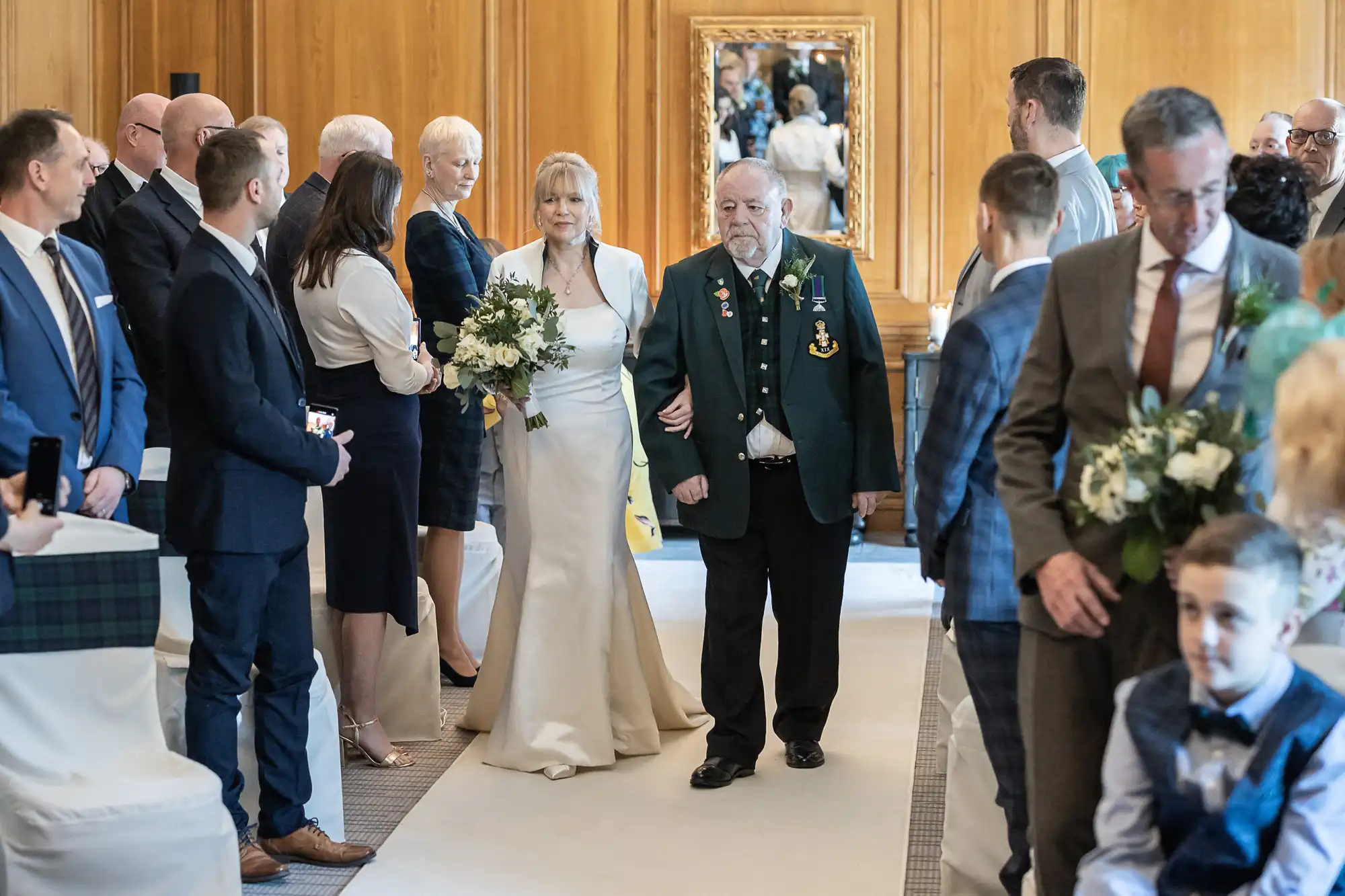 A bride in a white gown walks down the aisle with an elderly man wearing a green jacket adorned with medals, surrounded by standing guests in formal attire.