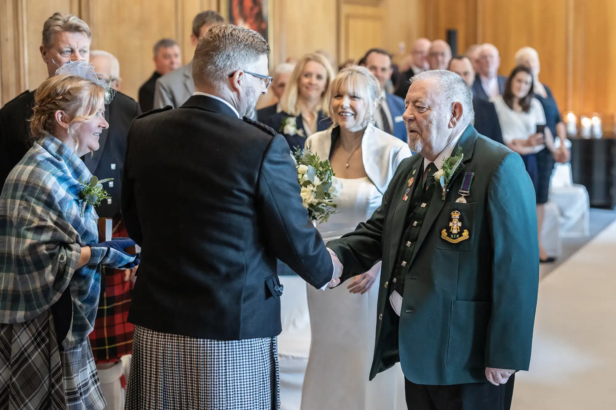 A groom in formal attire shakes hands with an older man in a green jacket at a wedding ceremony while other guests look on and smile.