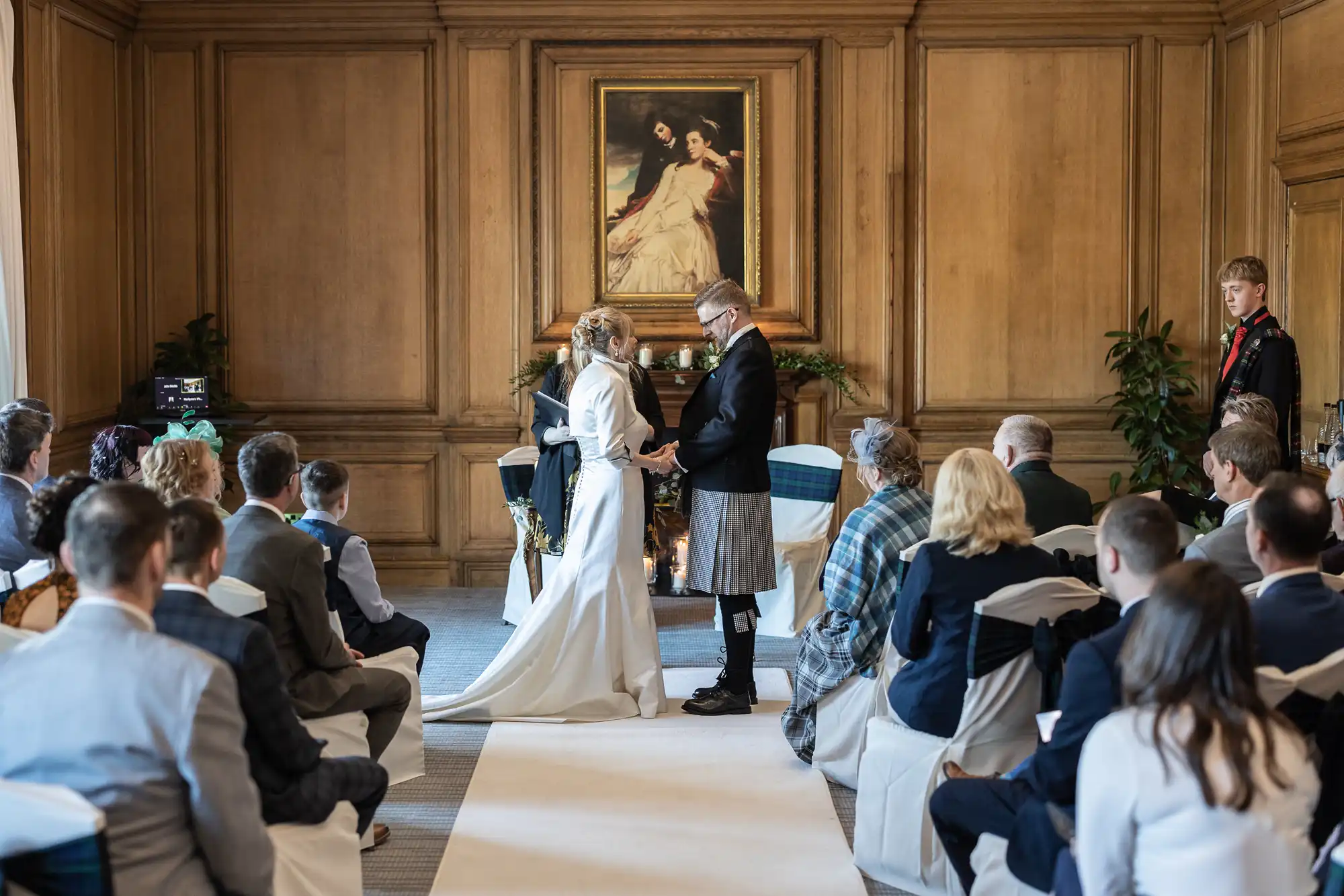 A couple stands in front of a registrar exchanging vows in a wood-paneled room, observed by seated guests. A large framed portrait is visible in the background.