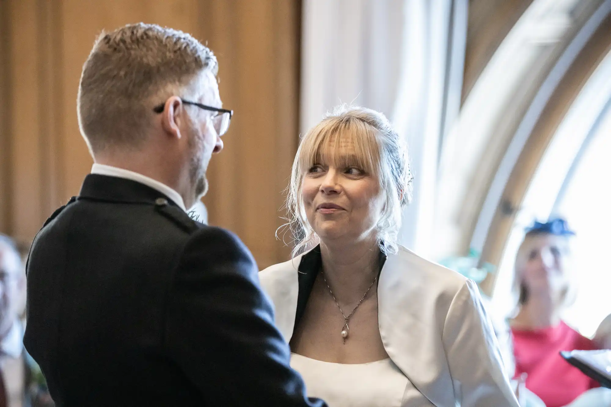 A man and a woman in formal attire stand facing each other, the woman smiling. An arched window and other guests are visible in the background.