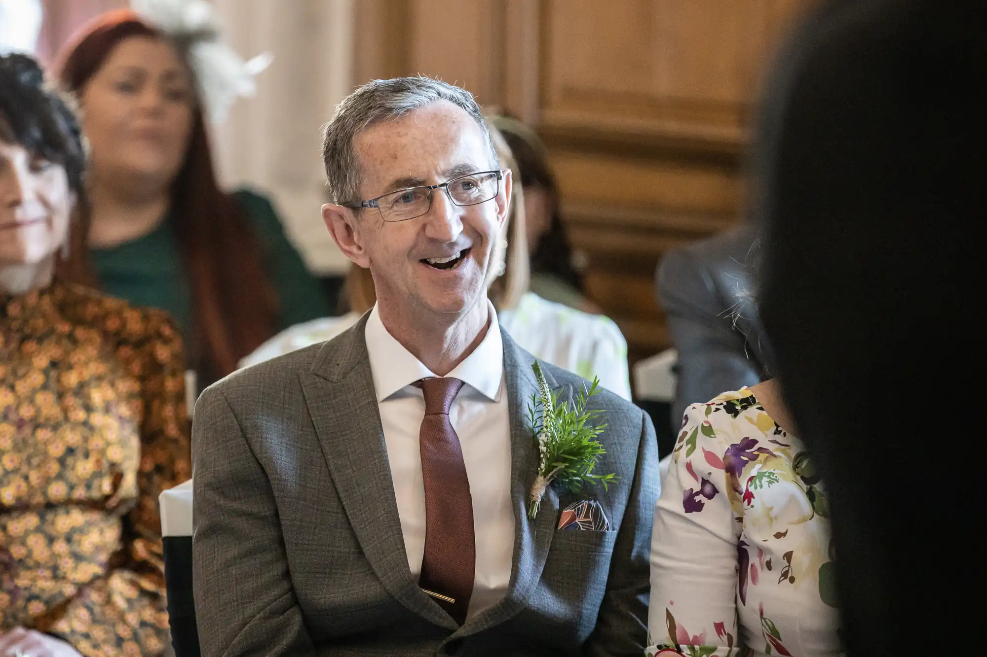 An elderly man in a suit and tie is smiling while sitting among a group of people in a room.