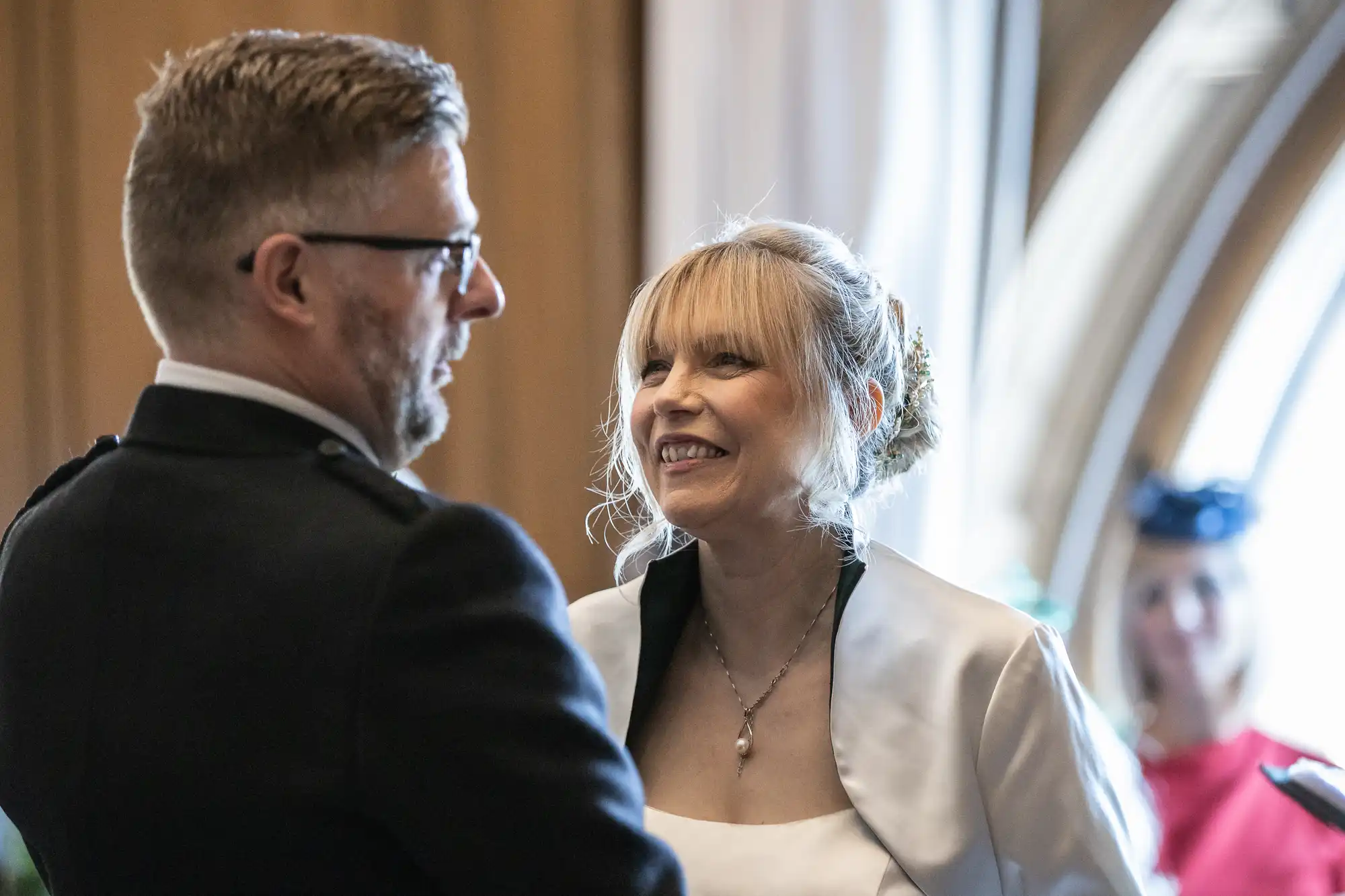 A man and a woman, both in formal attire, smile and exchange looks in a well-lit room with wooden accents and arched windows in the background.