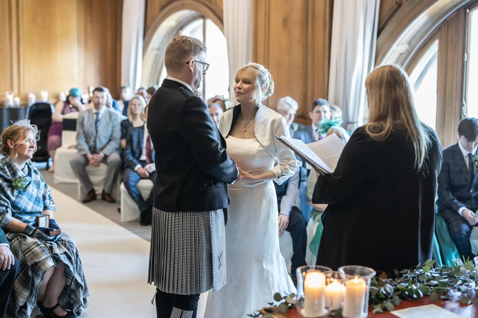 A couple stands holding hands during a wedding ceremony, officiated by a woman reading from a book. Guests are seated, watching the ceremony in a wood-paneled room with arched windows.