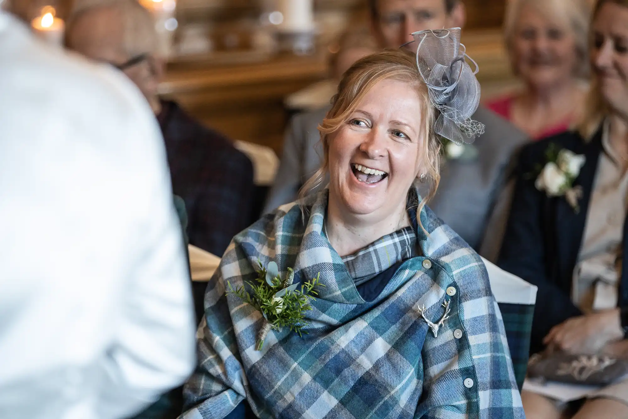 A woman in a plaid outfit and fascinator laughs while sitting among other attendees at a formal event.