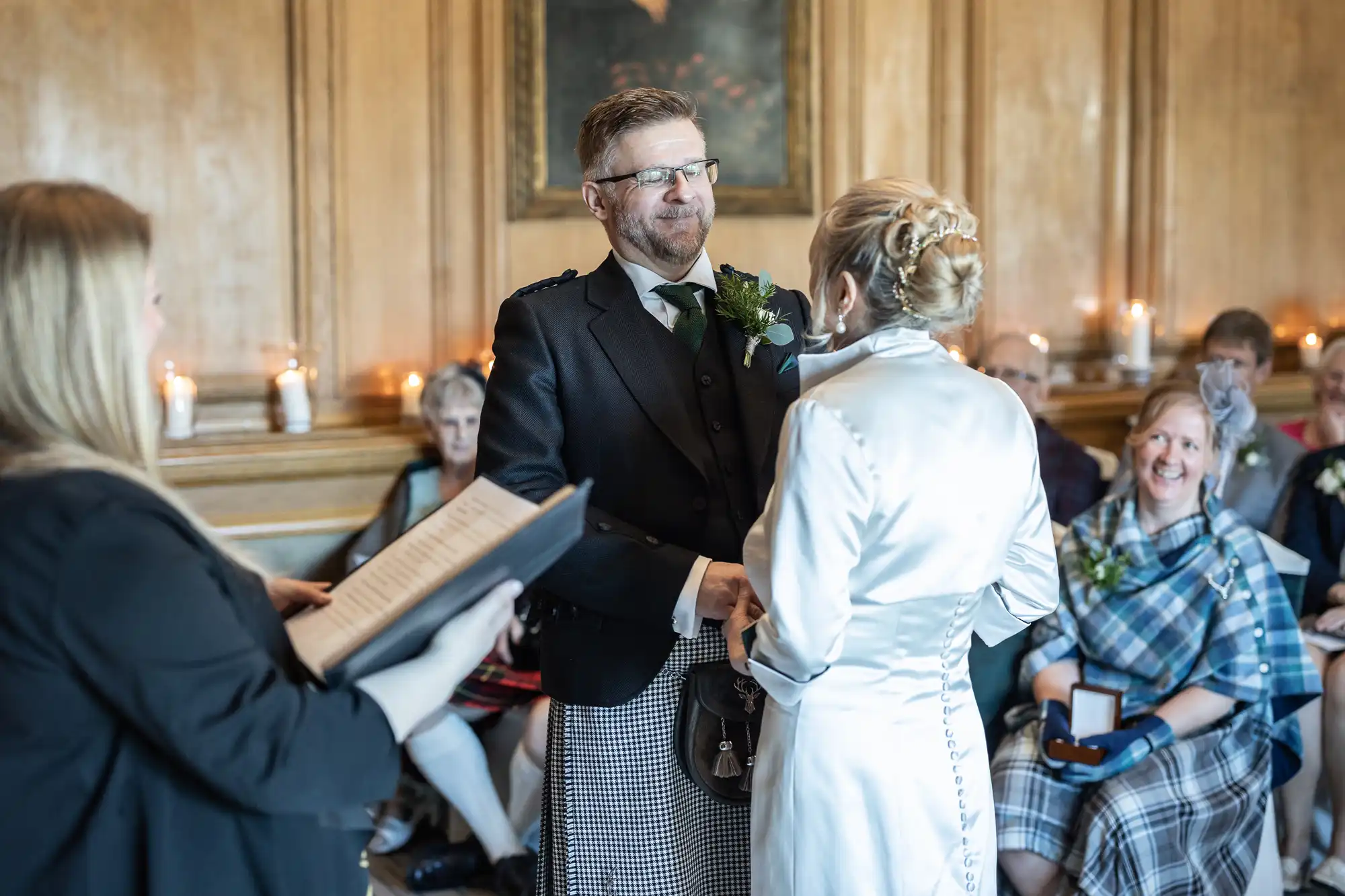 A couple stands facing each other, holding hands, during a wedding ceremony. A person on the left reads from an open book, with seated guests watching in the background.