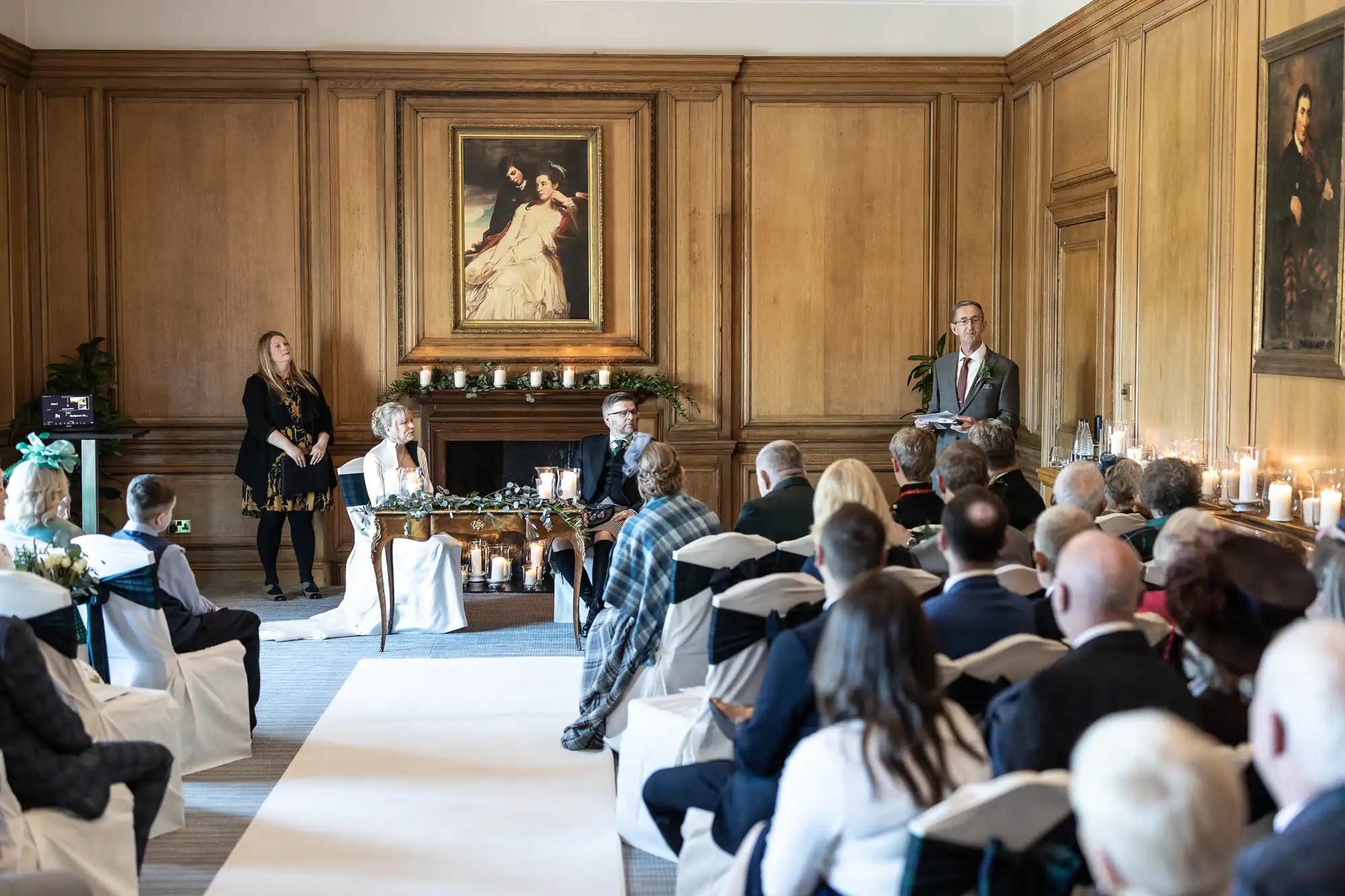 A wedding ceremony in a wood-paneled room with guests seated, a couple at the front, and officiants standing by.