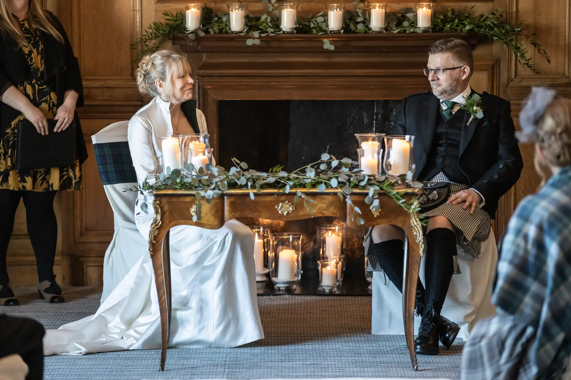 A bride and groom are seated at a small table adorned with candles and greenery in front of a fireplace, during a wedding ceremony. The bride wears a white dress, and the groom wears a black suit with a kilt.