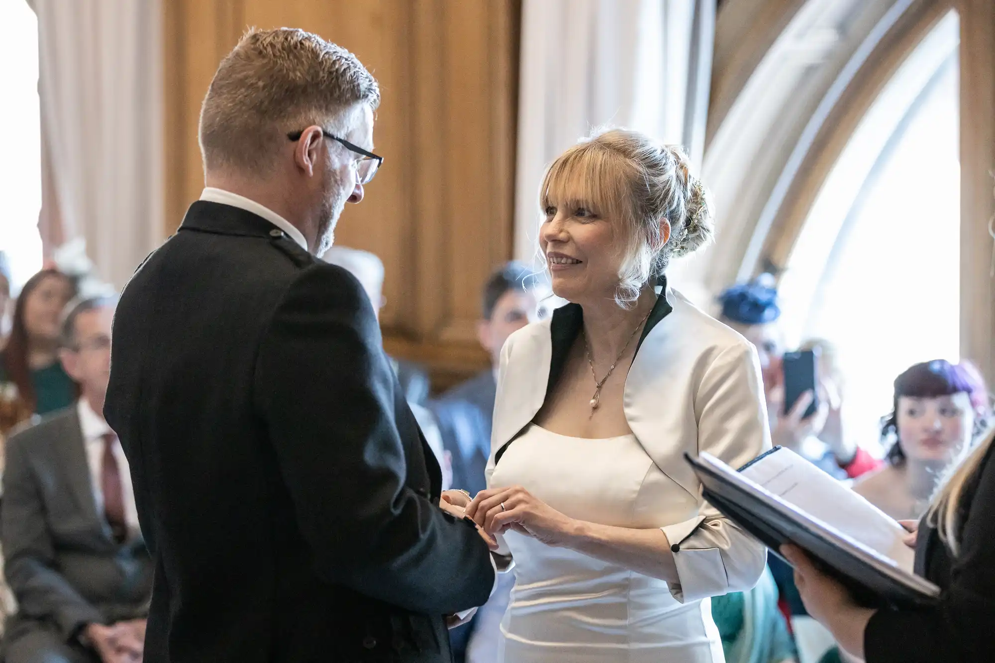 A couple stands facing each other, holding hands and exchanging rings during a wedding ceremony. The bride wears a white dress and the groom is dressed in a dark suit. Guests are seated in the background.