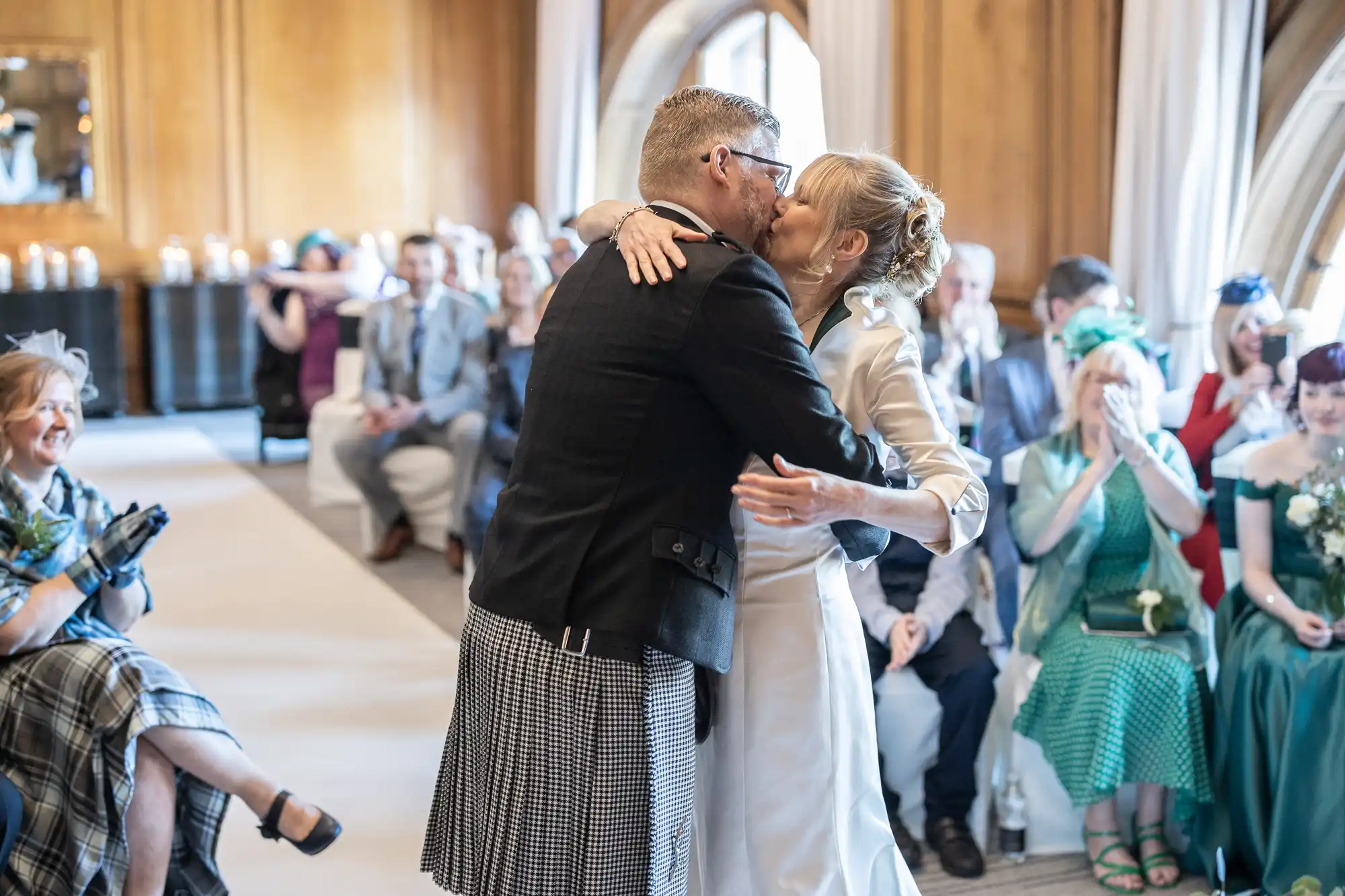 A couple kisses during their wedding ceremony in a room filled with seated guests. The groom is wearing a traditional Scottish kilt, and the bride is dressed in white.