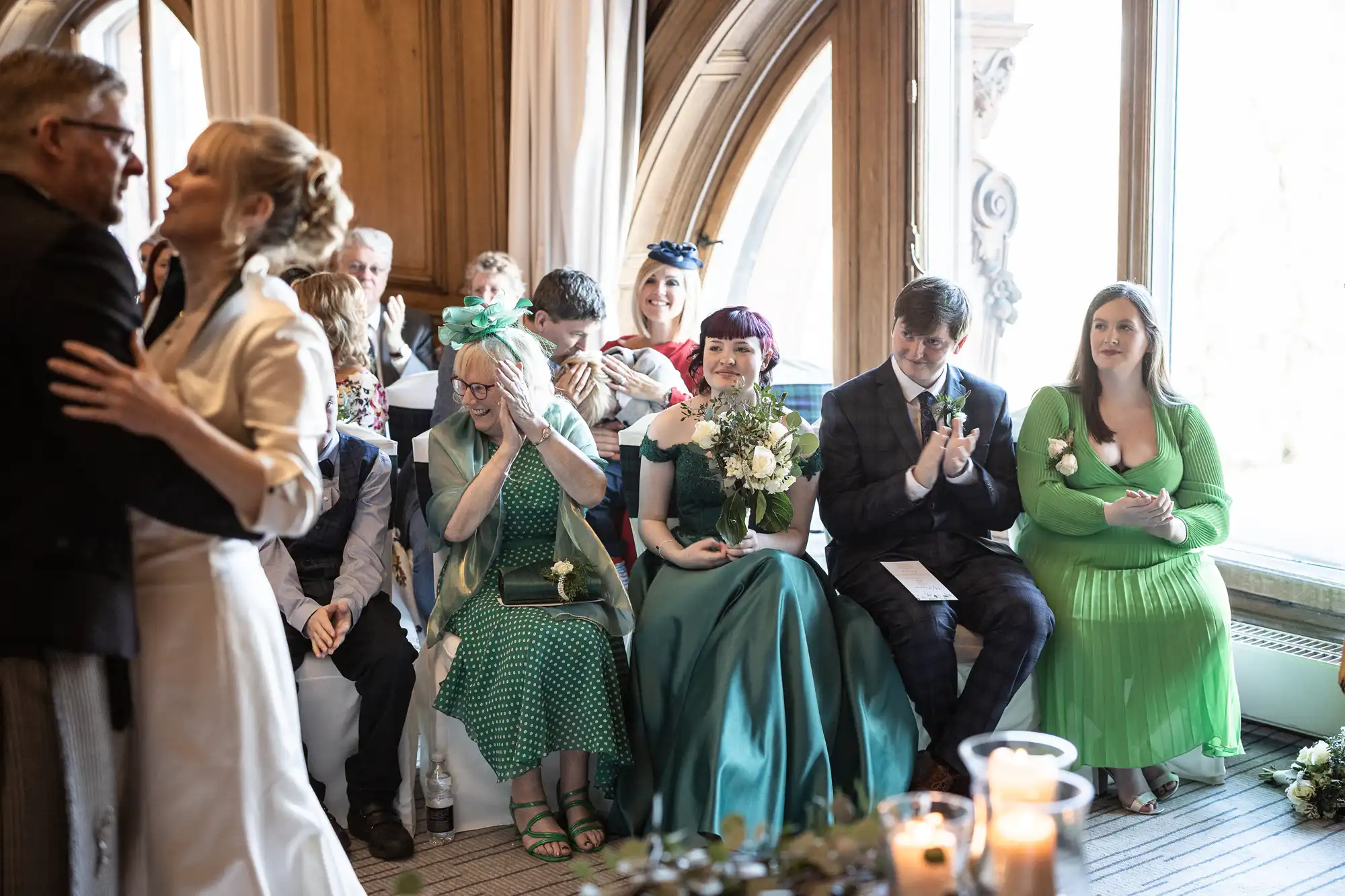 A couple dances in front of an audience at a formal event. Attendees are dressed in formal and semi-formal attire, with some clapping and others watching attentively. Candles are visible in the foreground.