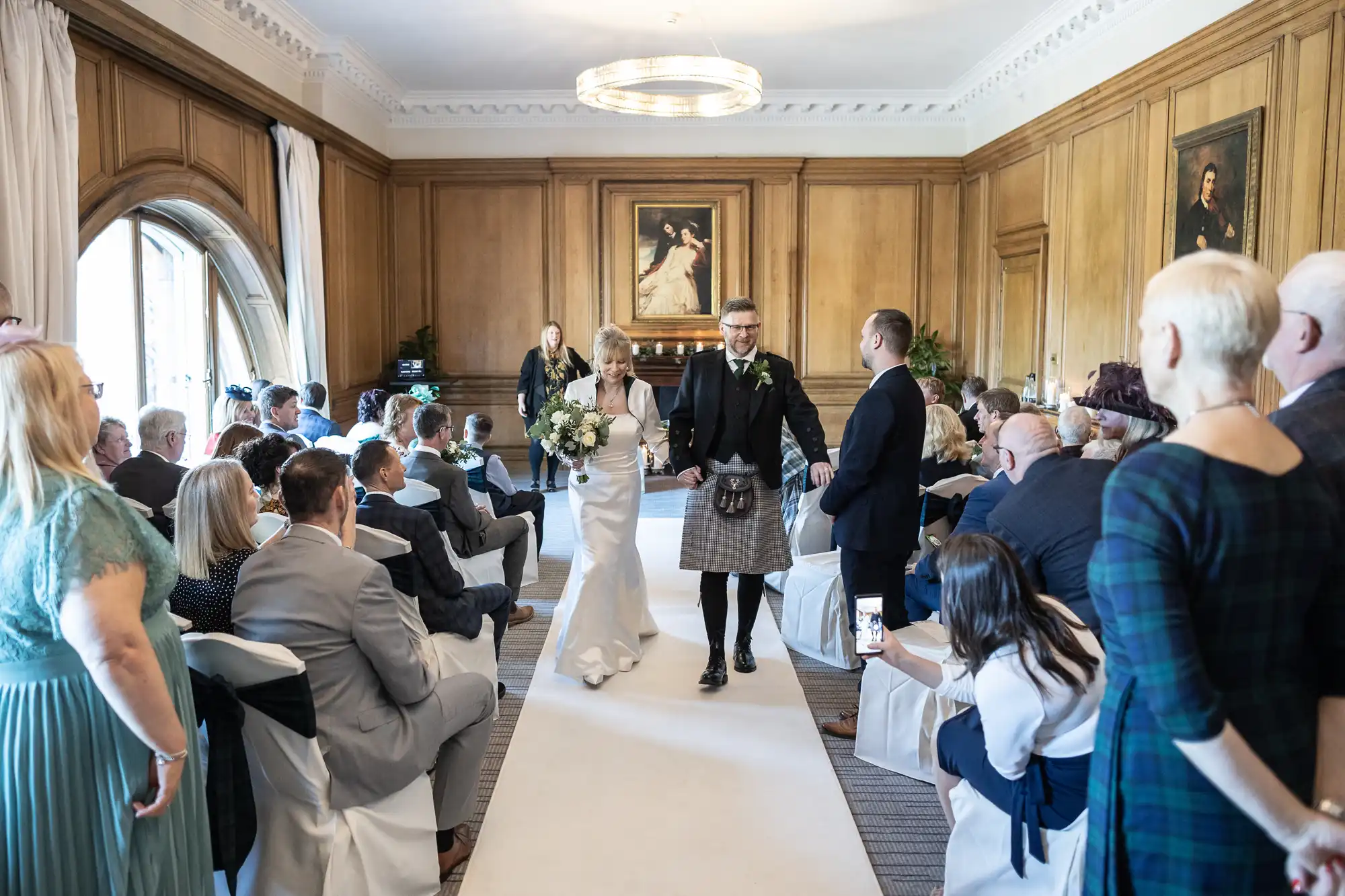 A bride and groom walk down the aisle of a wood-paneled room full of seated guests during their wedding ceremony.