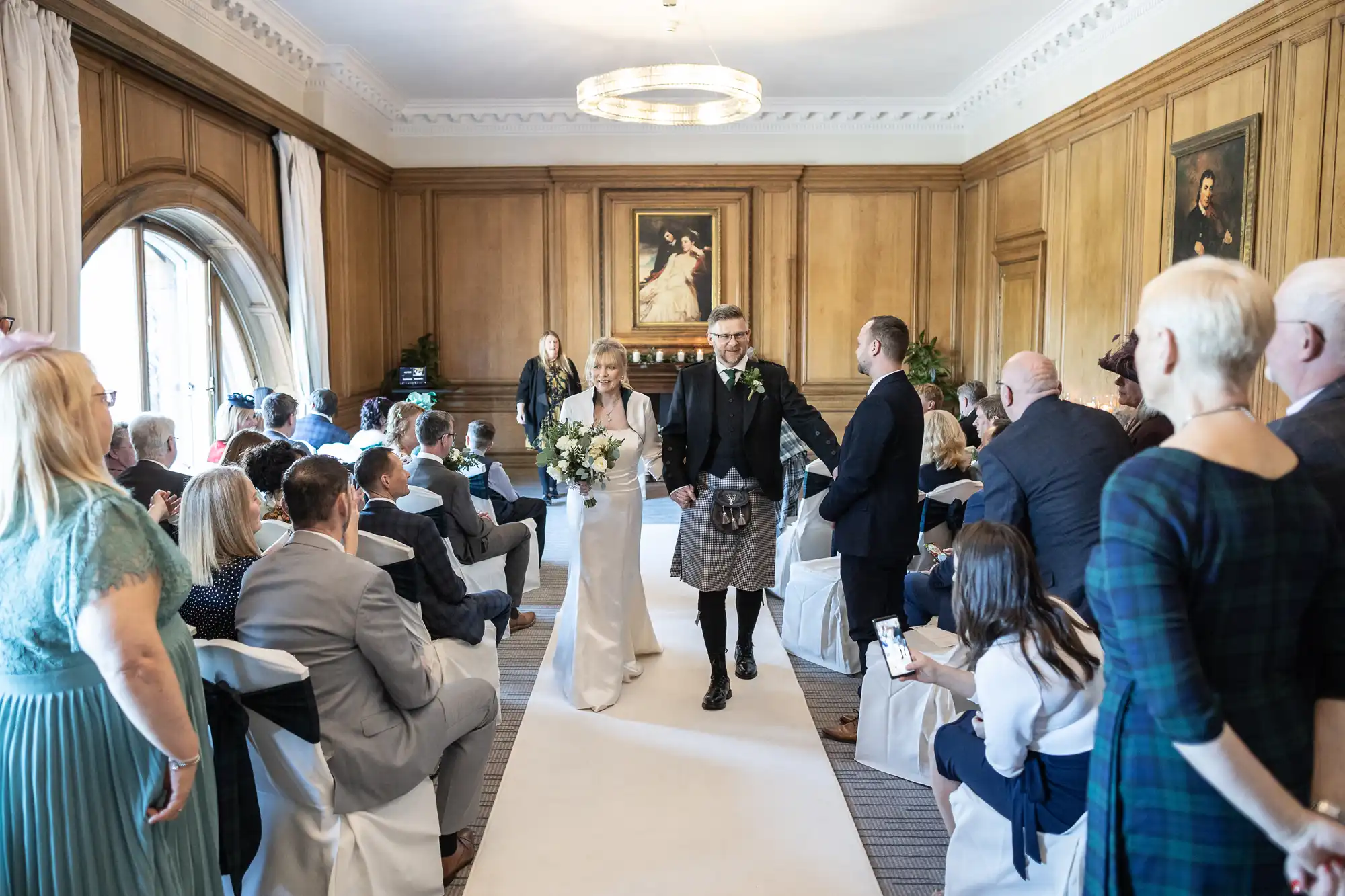 A bride and groom walk down the aisle in a wood-paneled room, with seated guests on either side and a chandelier overhead.