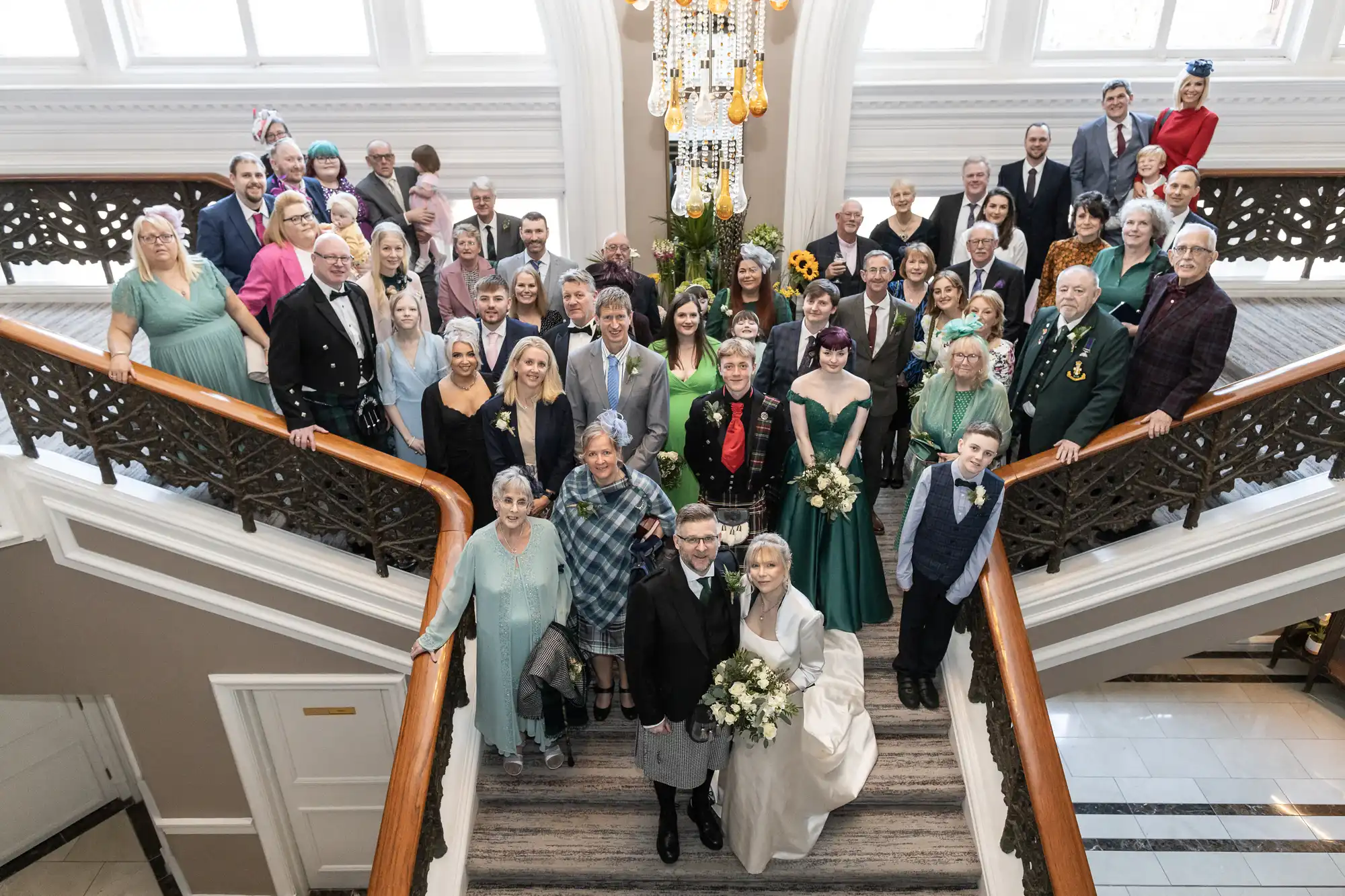 A large wedding group photo taken on a staircase with the bride and groom at the base, surrounded by family and friends. Elegant interior setting with bright lighting and chandelier overhead.