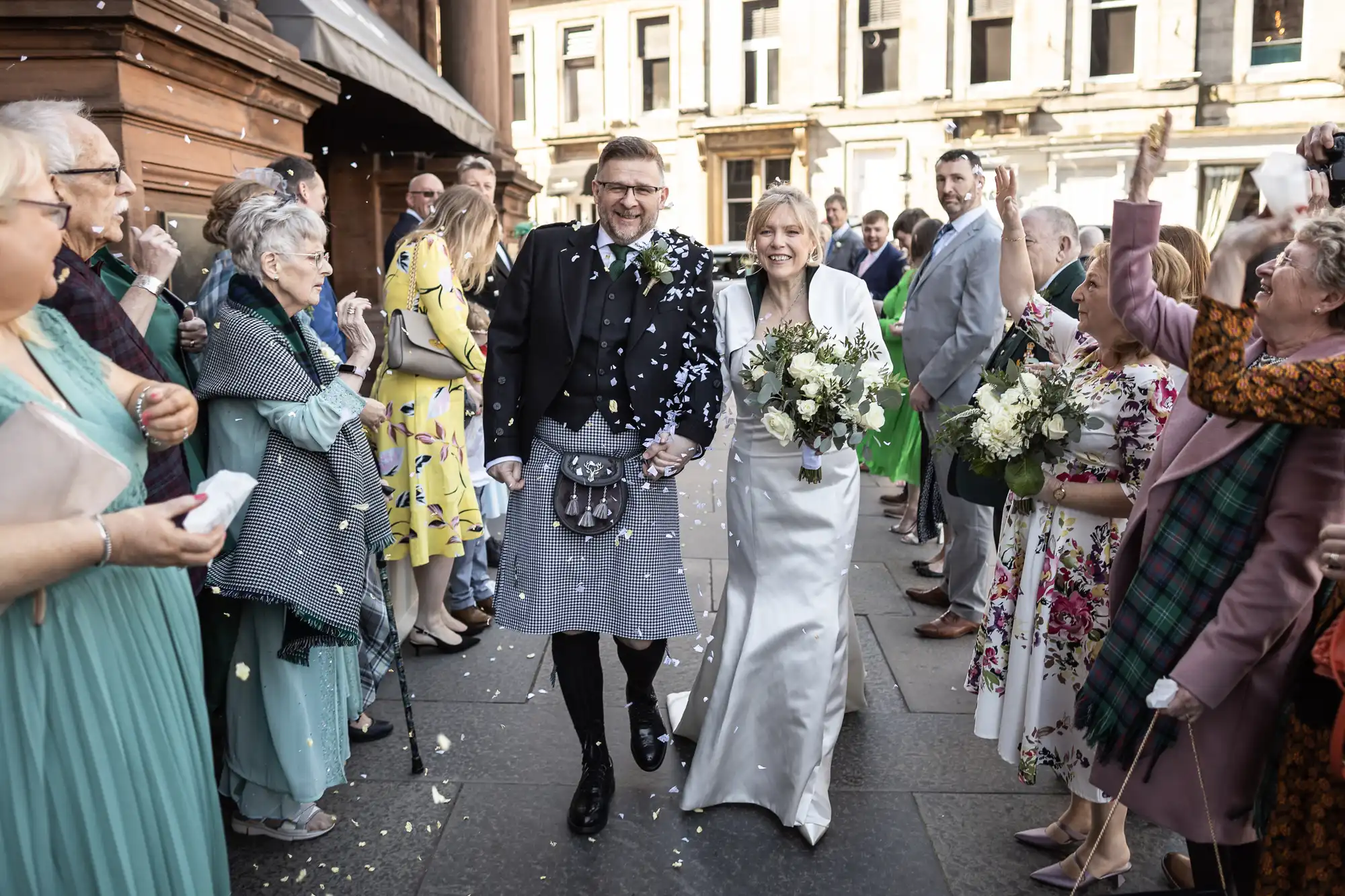 A bride and groom, both smiling and dressed in traditional wedding attire, walk hand-in-hand down a stone path while guests on either side throw confetti and celebrate.