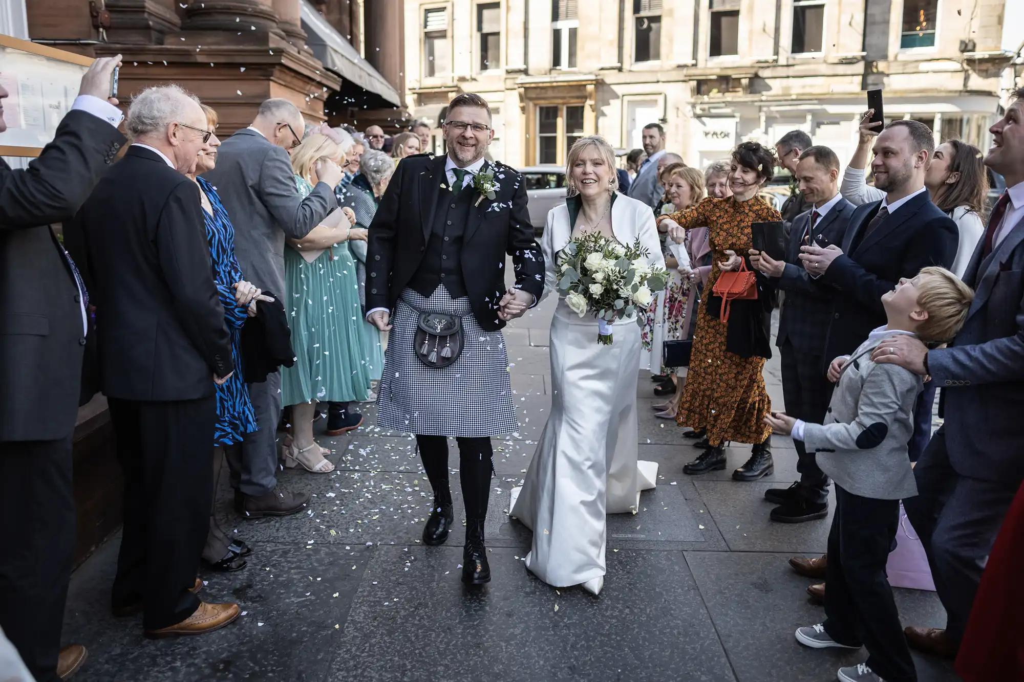 A bride and groom walk down an aisle outdoors, surrounded by guests who are clapping and throwing confetti. The bride is holding a bouquet and wearing a white dress; the groom is in a suit with a kilt.