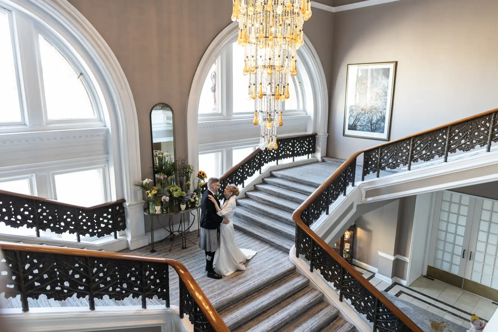 A couple stands on a grand staircase under an ornate chandelier in an elegant venue with arched windows and decorative railings.