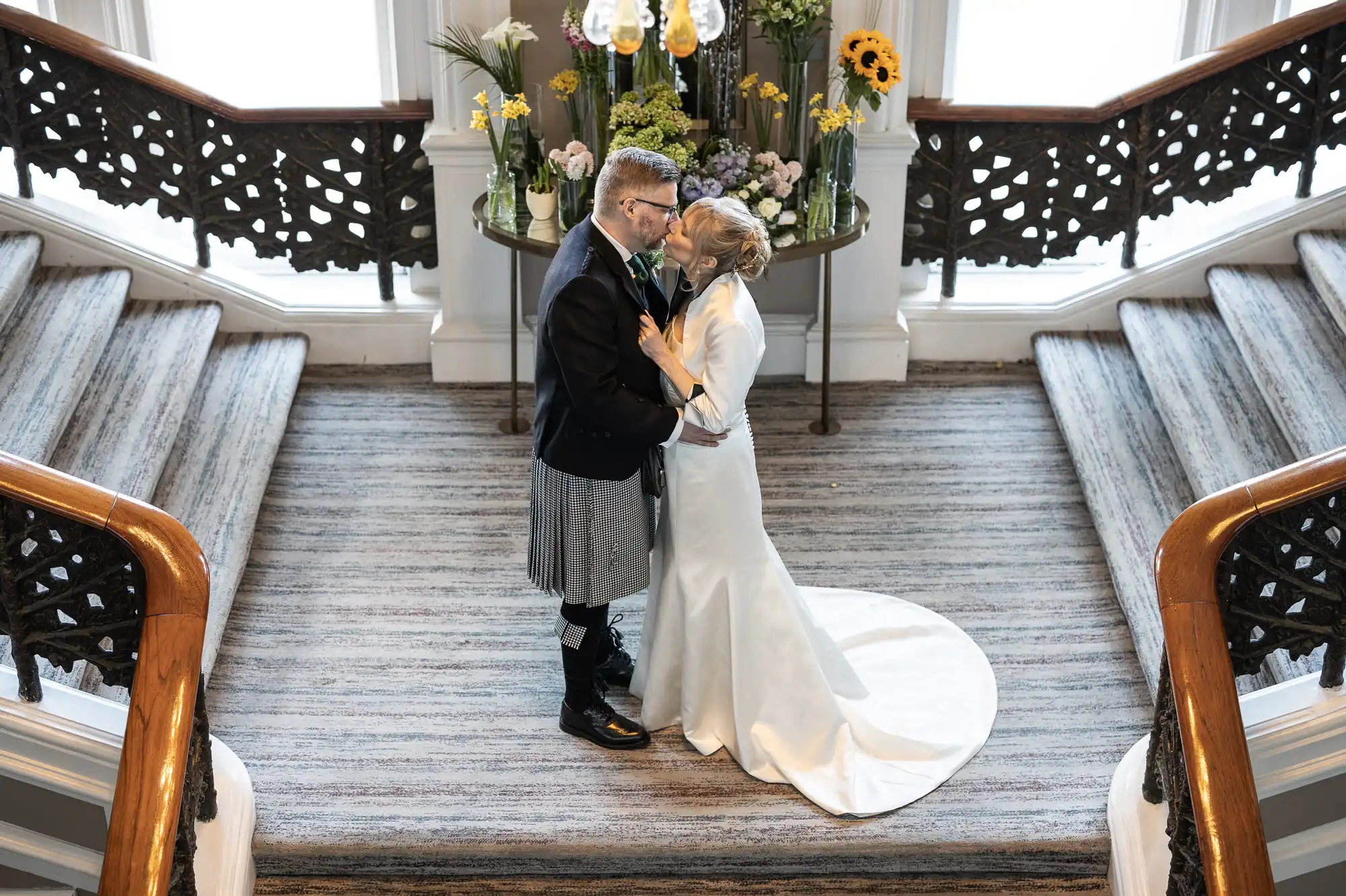 A wedding couple embraces at the bottom of a split staircase in a formal setting with flowers on a table at the background. The groom wears a kilt while the bride is in a white gown.