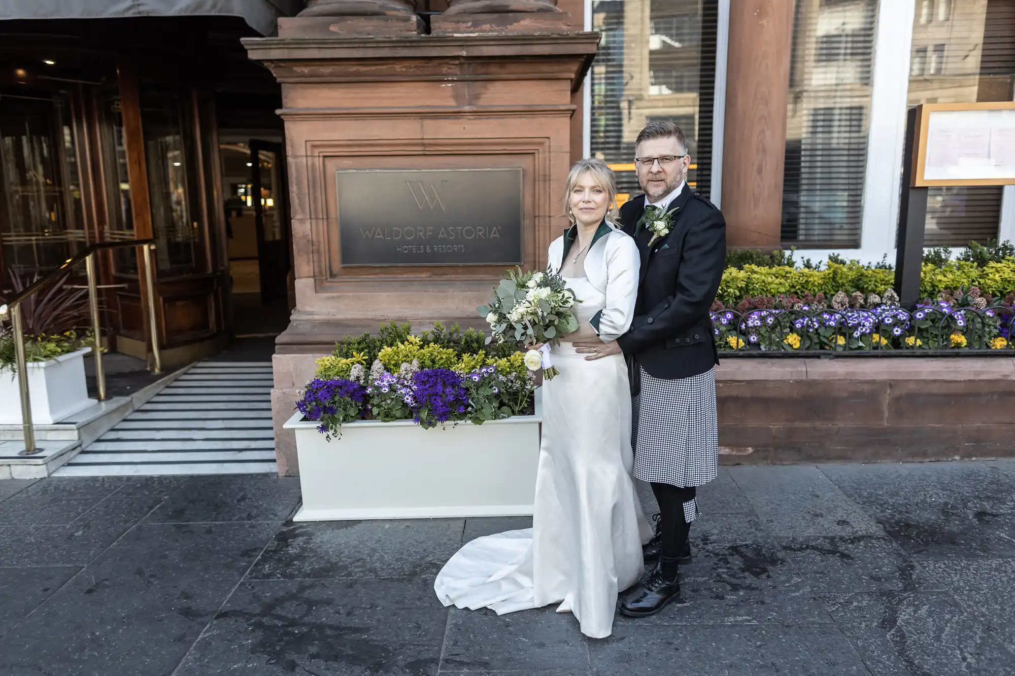 A couple poses outside the Waldorf Astoria, with the woman in a white gown holding a bouquet and the man in traditional formal wear. Flower boxes are placed by the doorway.