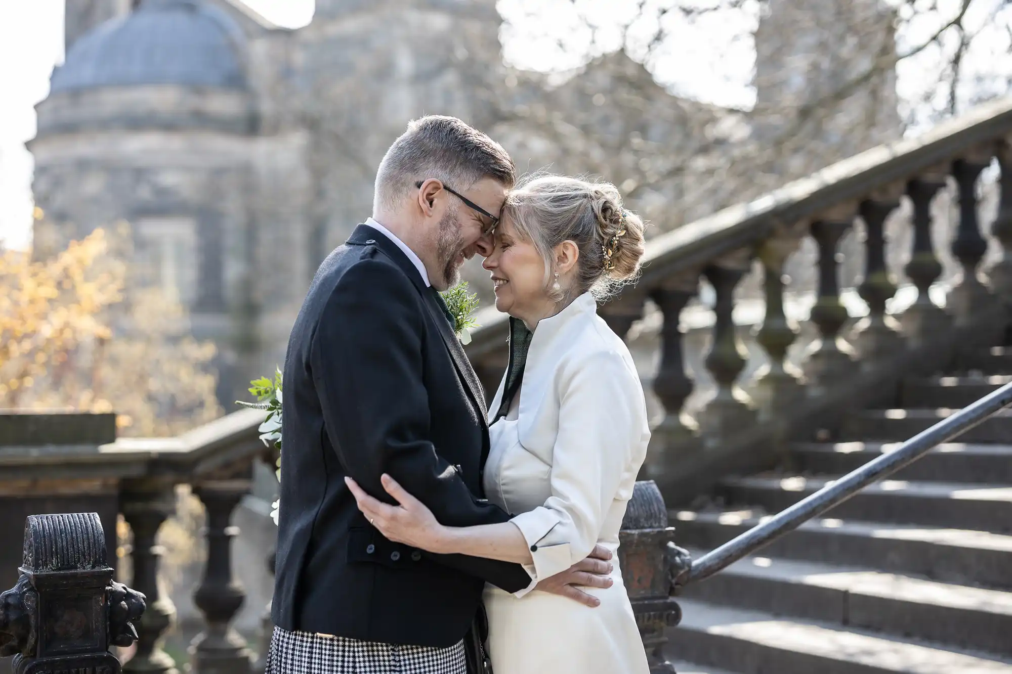 A couple in formal attire embraces and smiles at each other on the steps of an outdoor stone staircase with historic buildings in the background.