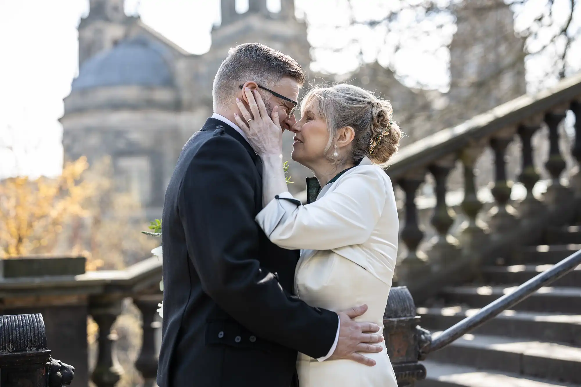 A couple dressed in formal attire shares a tender moment on an outdoor staircase with historic architecture in the background.