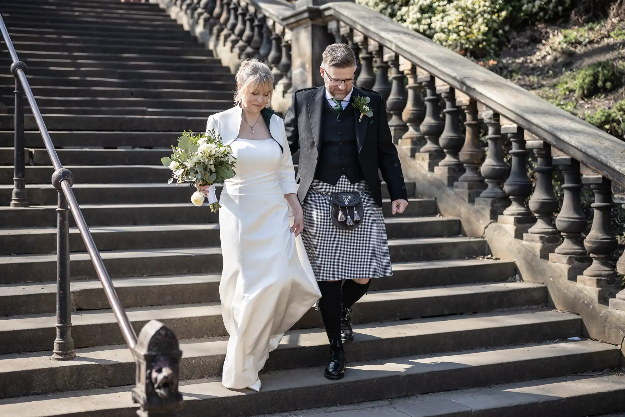 A couple dressed in wedding attire walks down stone stairs; the bride holds a bouquet and the groom wears a kilt, both descending side by side.