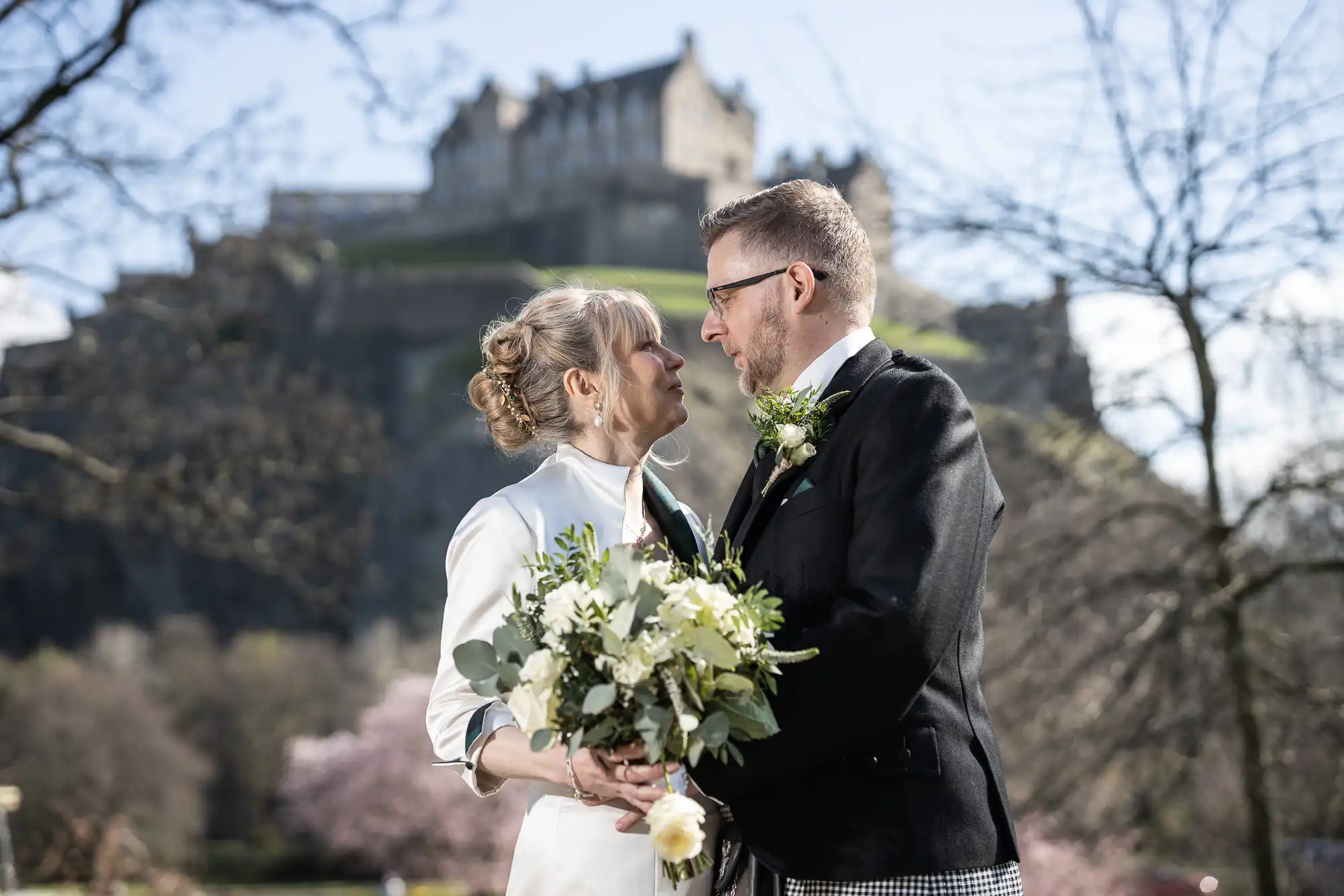 A couple in wedding attire stands close, holding a bouquet, with a historic castle on a hill in the background.