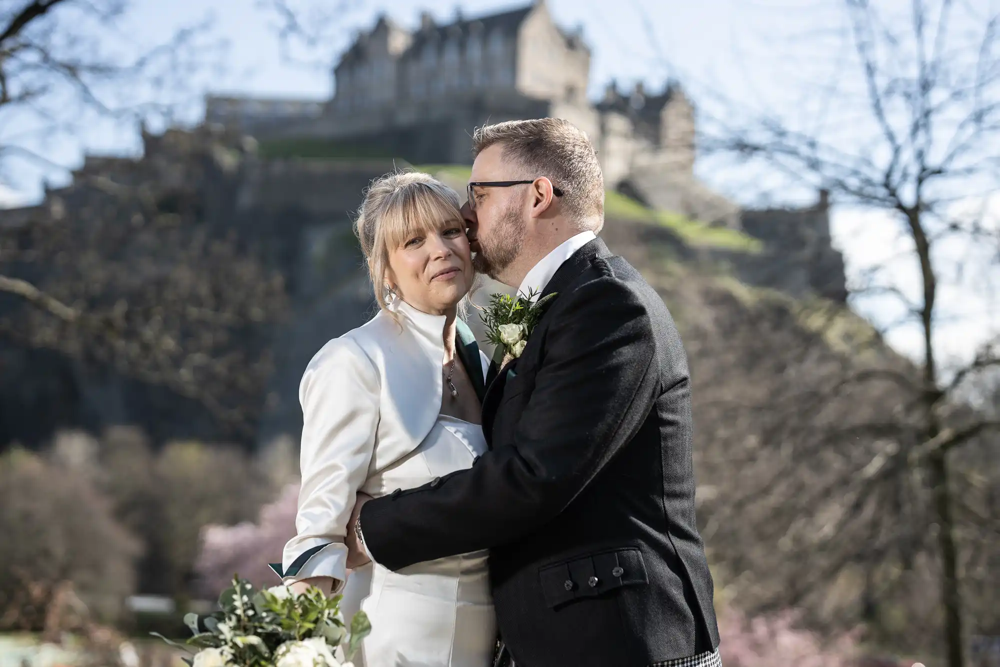 A couple in formal attire, with the man kissing the woman's cheek, stands outdoors in front of a historic building on a clear day.