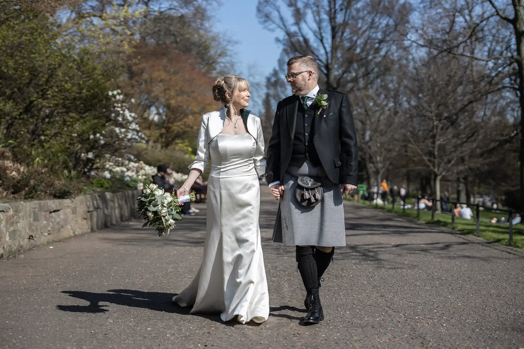 wedding photographer at the Caledonian Hotel: a bride in a white satin dress and groom in a kilt walk hand-in-hand along a park path on a sunny day.