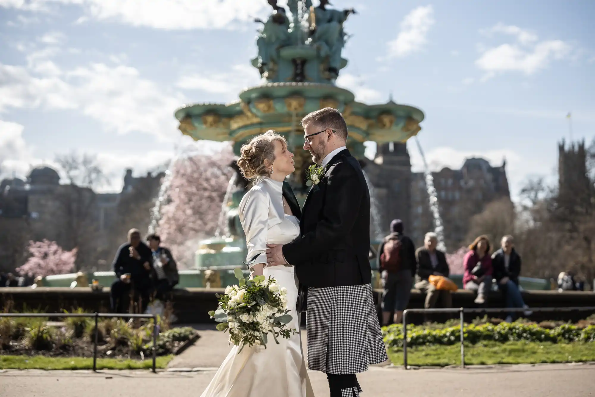 A bride and groom stand facing each other in front of a fountain, holding hands and smiling, with several people sitting on a bench in the background.