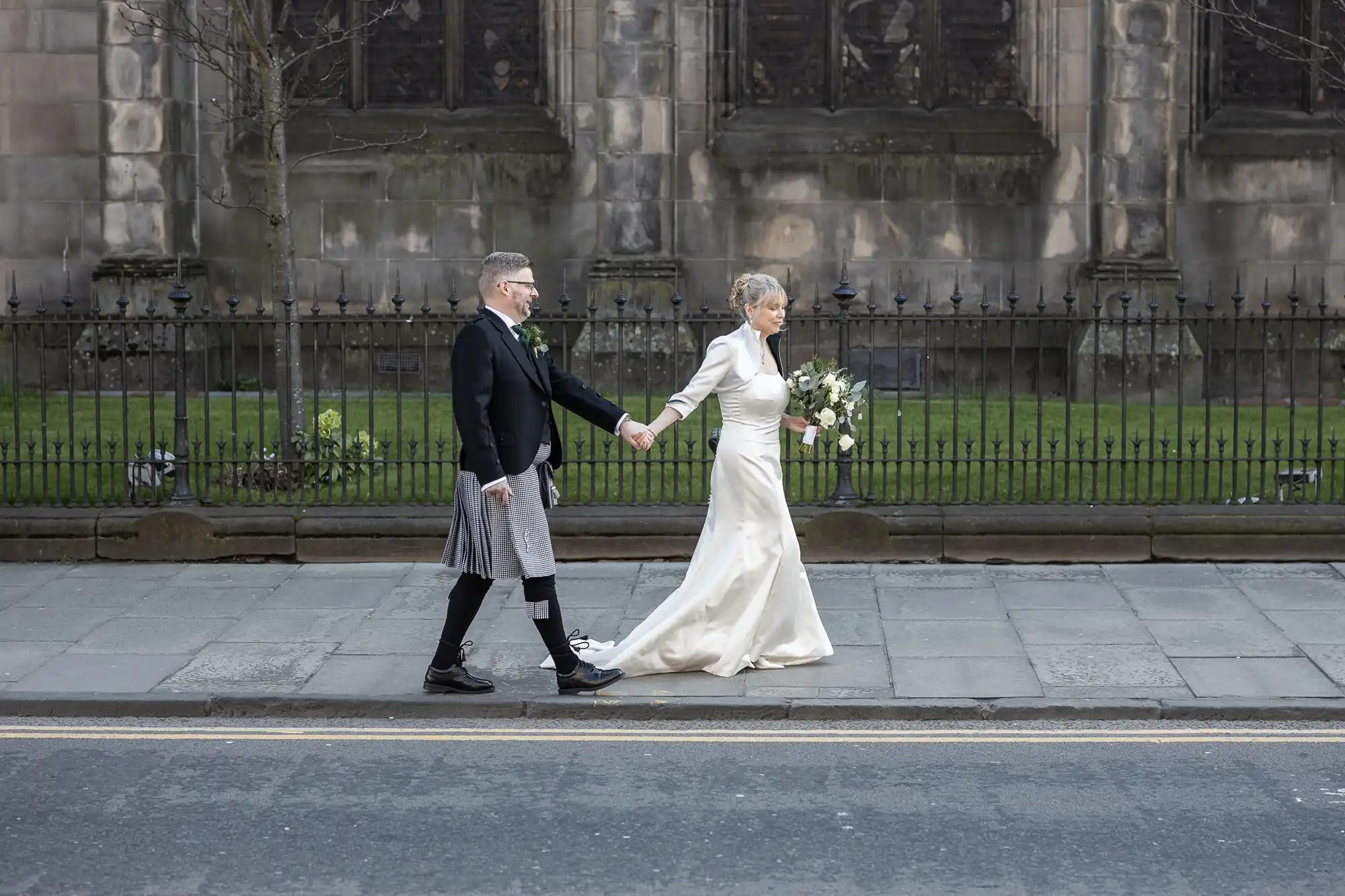 A couple in wedding attire, with the groom in a kilt and the bride in a white gown, walk hand in hand on a sidewalk in front of an old stone building.