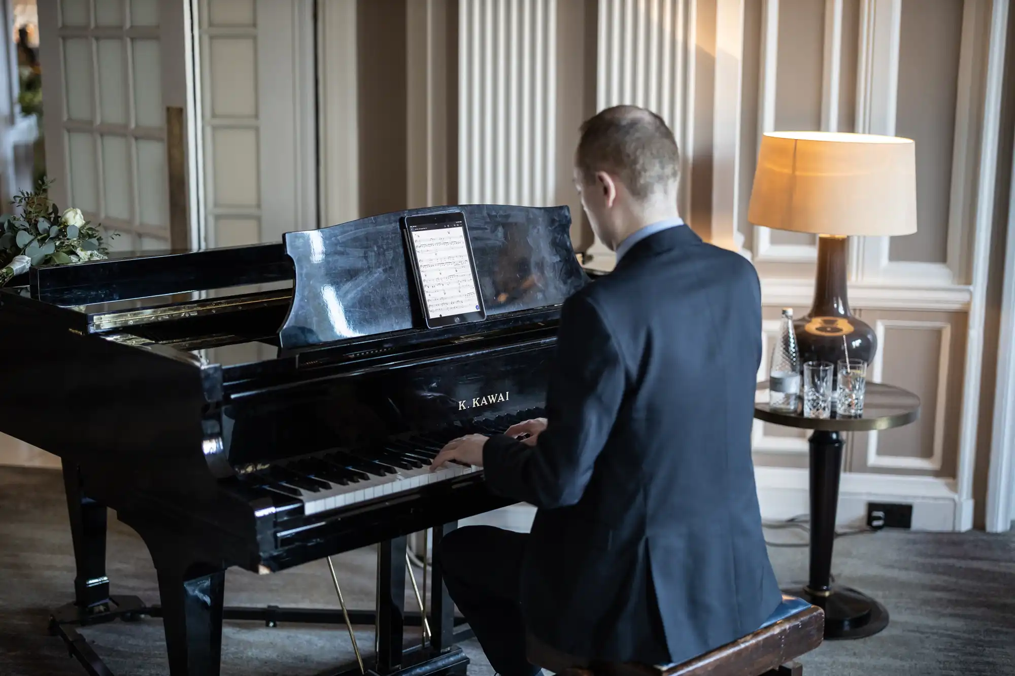 A person in a suit plays a black grand piano in a well-lit room, with a tablet displaying sheet music on the music stand.