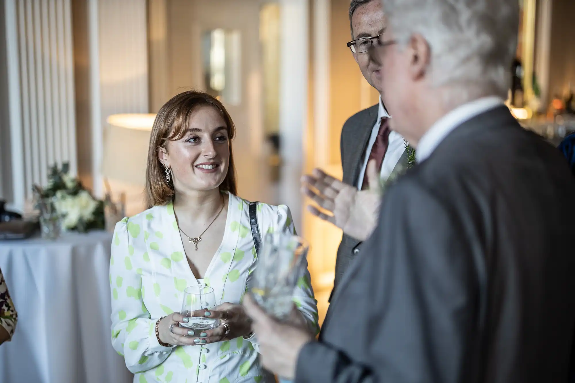 A woman in a white polka-dot dress stands in conversation with two older men in suits at a social event. One man holds a glass, and blurred details of the room and attendees are visible in the background.