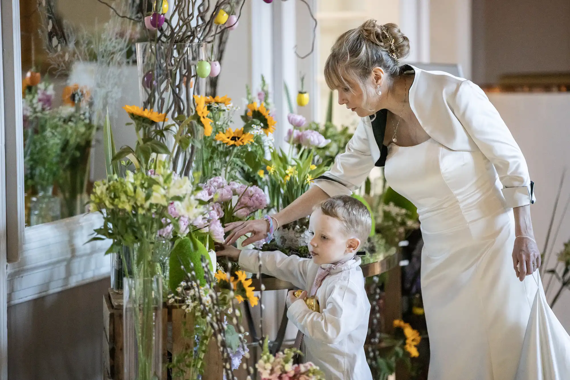 A woman in a white dress and a young boy in formal attire look at a variety of colorful flowers, with decorative branches hanging above.