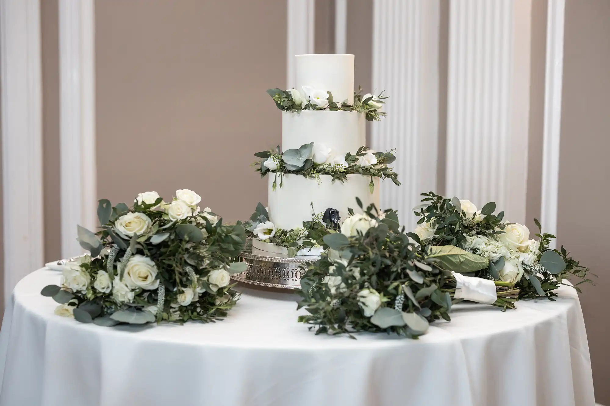 A three-tier white wedding cake decorated with green foliage and white flowers is placed on a table alongside four floral bouquets.