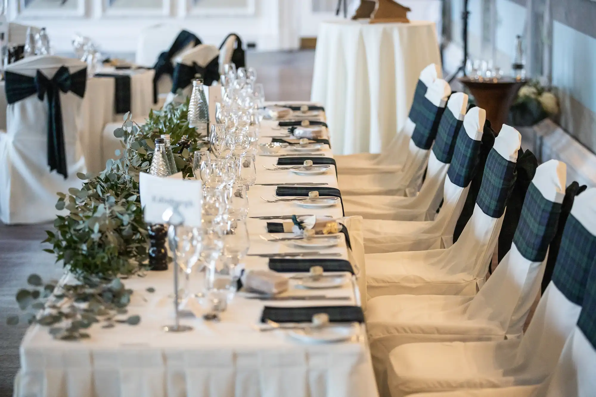 A formal dining table is set with white tablecloths, black and white chair covers, silverware, wine glasses, and greenery as a centerpiece. The setting is arranged for multiple guests.