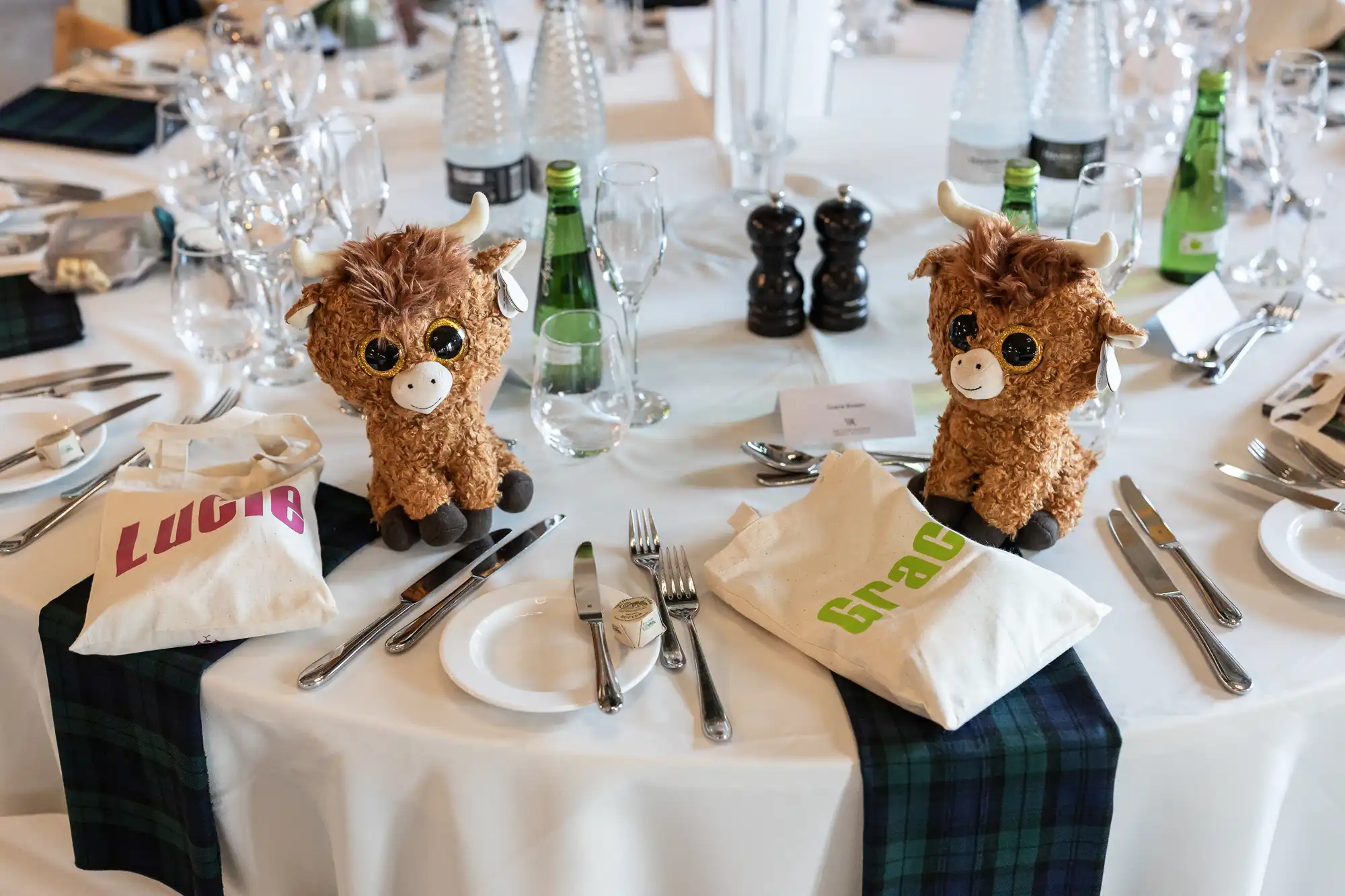 A round dining table set with dishes, cutlery, glasses, and bottles. Two plush toy cows sit on two of the plates, each with a personalized tote bag labeled "Lucía" and "Grace.