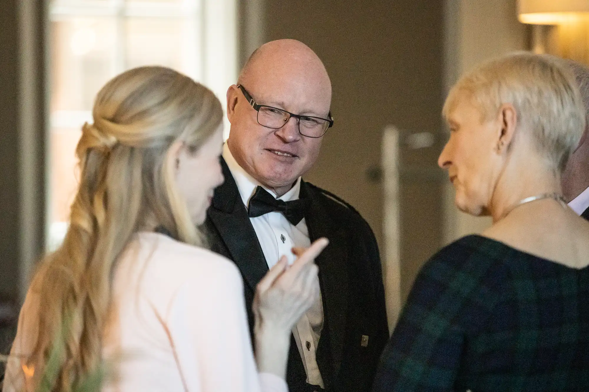 Three people are engaged in a conversation at a formal event. One man in a tuxedo is talking with two women; one has blonde hair and is wearing a light-colored top, and the other has short, blonde hair.