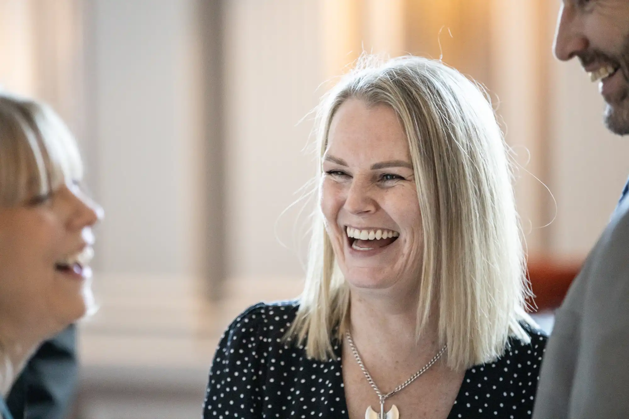 A woman with blonde hair and a black polka dot top is smiling and laughing in conversation with two people.