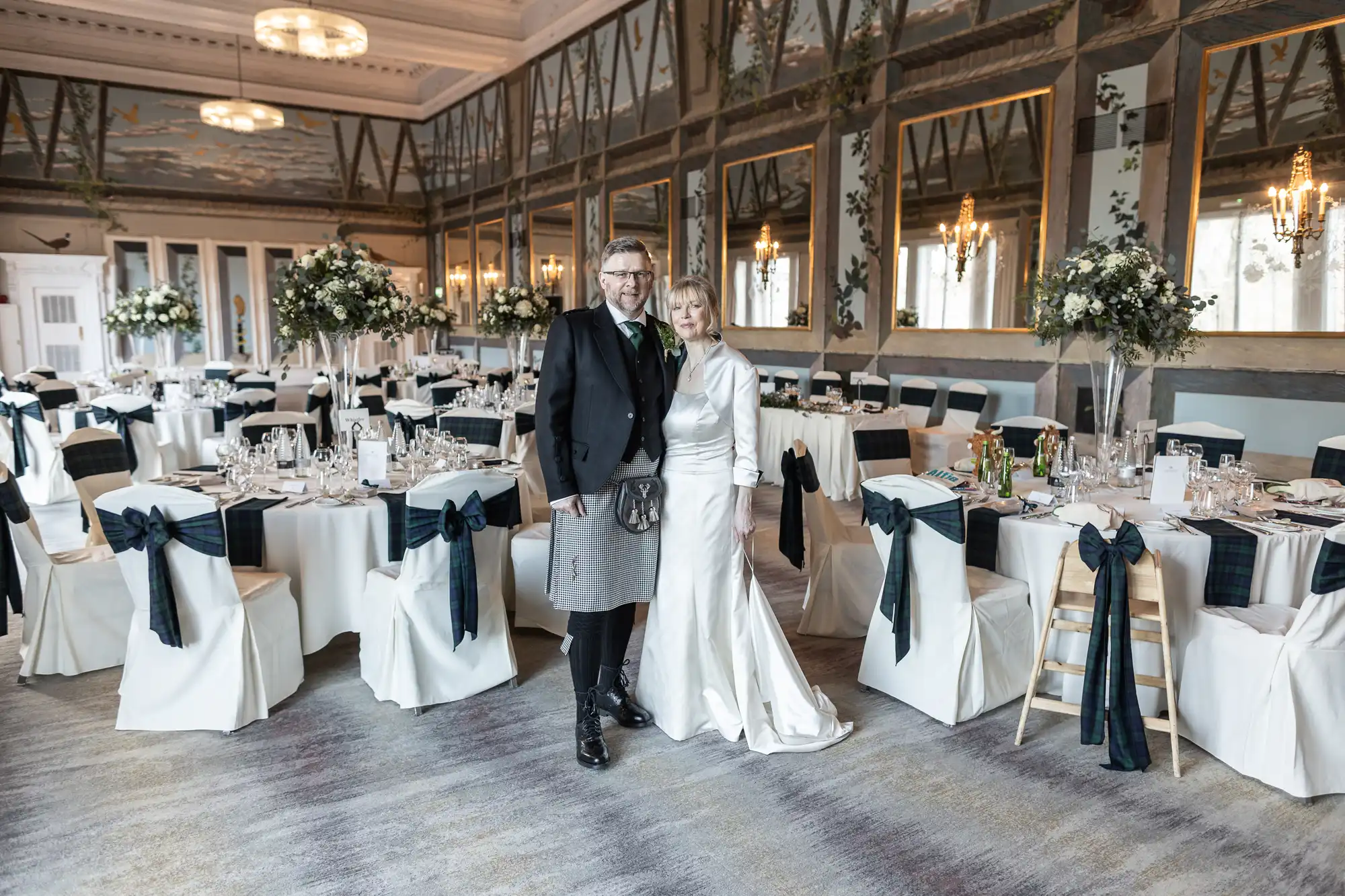 A couple stands in a decorated banquet hall with white and navy-accented chairs, tables set for dining, and chandeliers hanging from the ceiling.