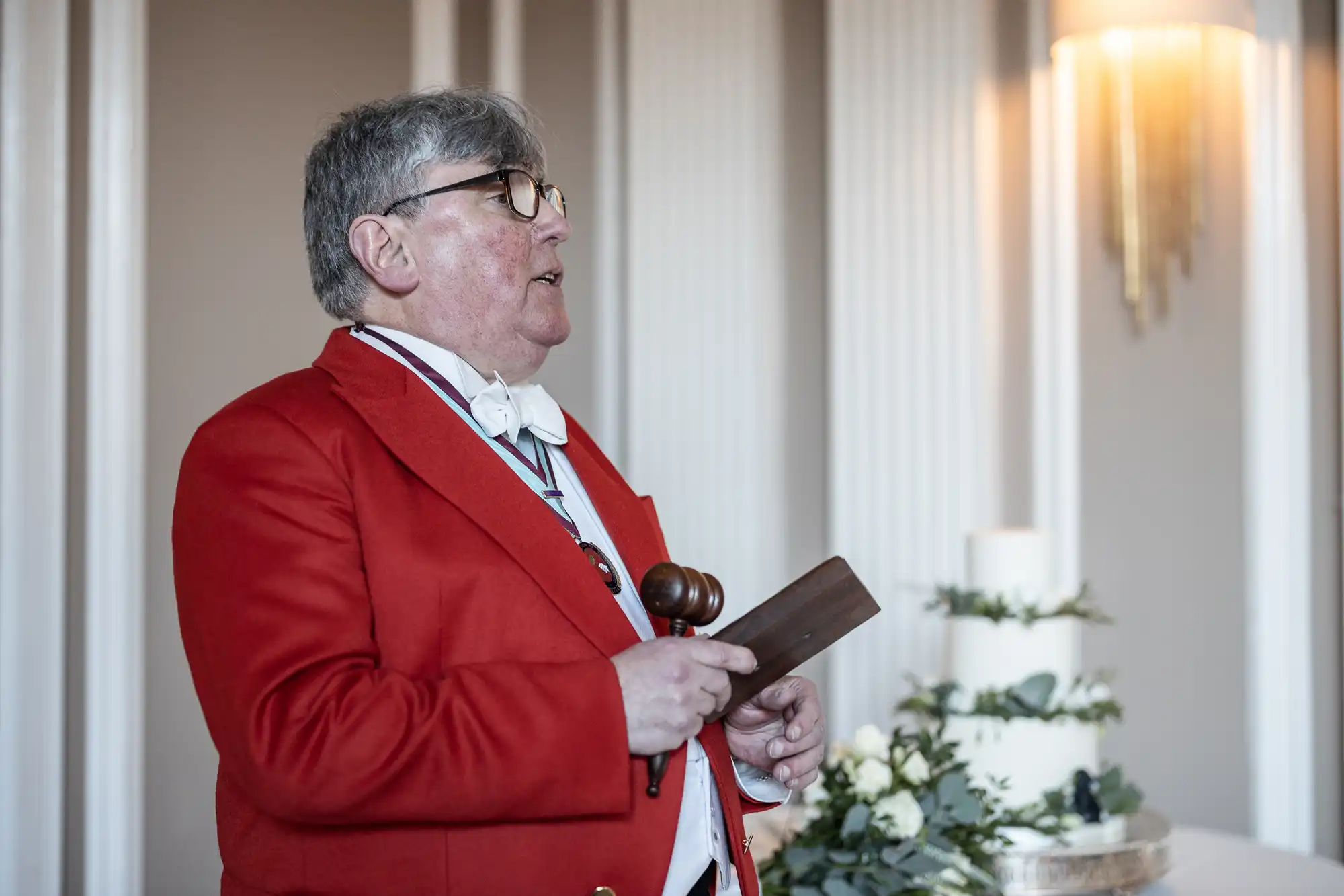 Person in a red jacket holding a gavel, standing near a table with a white tiered cake.