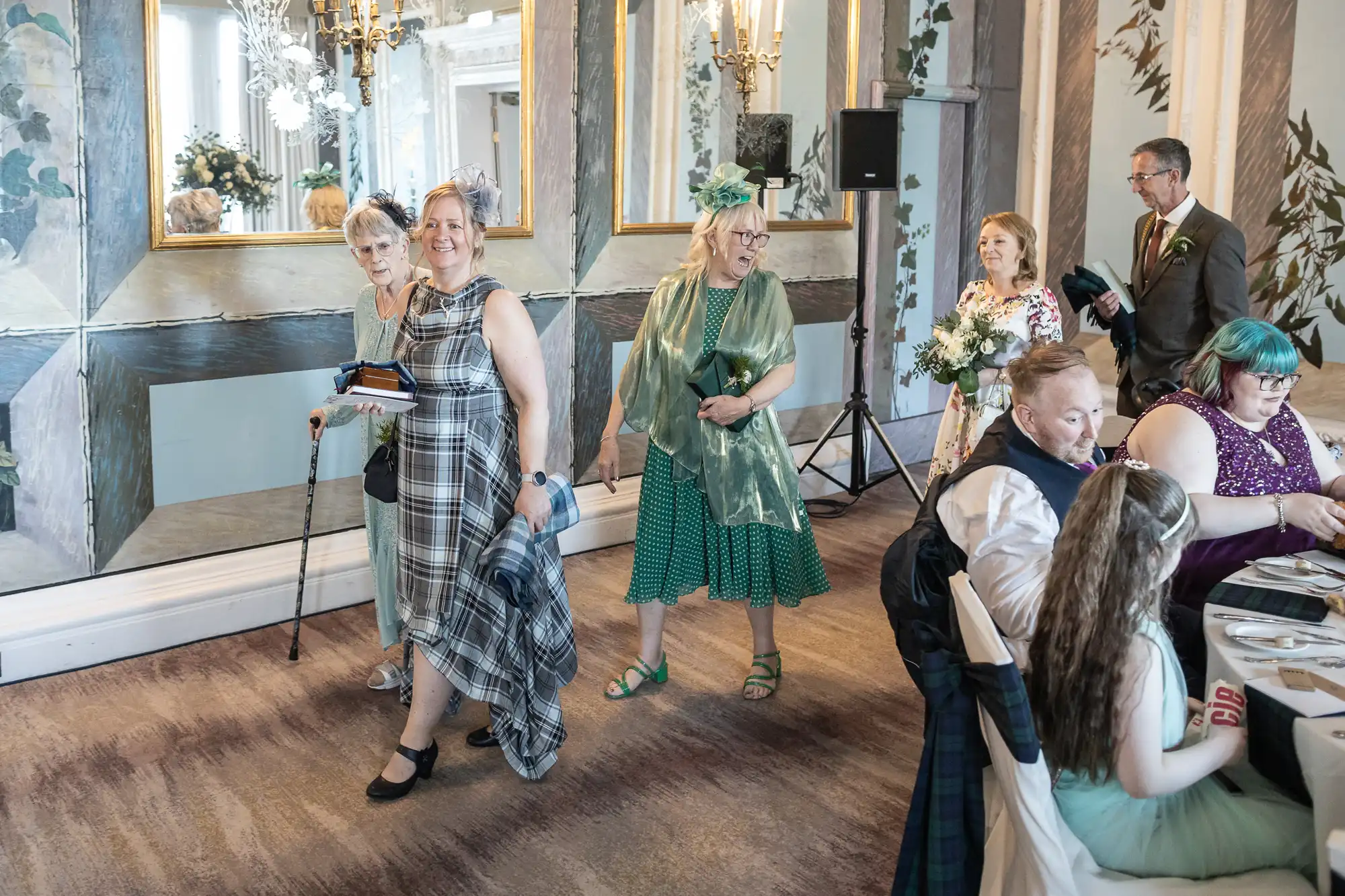 A group of people, some holding bouquets and wearing various dresses, walk through a room decorated with large mirrors and chandeliers while others are seated at tables.